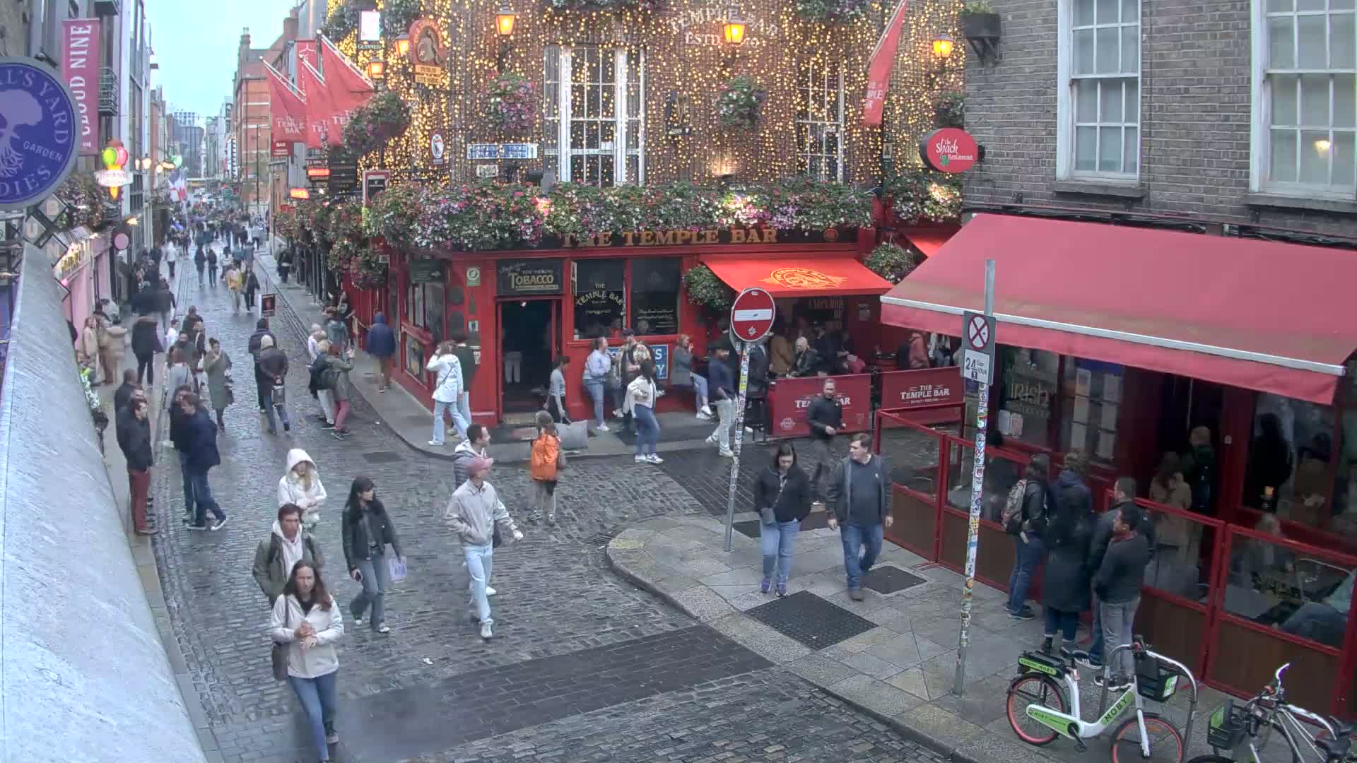A cobblestone street, possibly in a European city, is bustling with pedestrians on an overcast day, centered on a red building with string lights and outdoor seating.