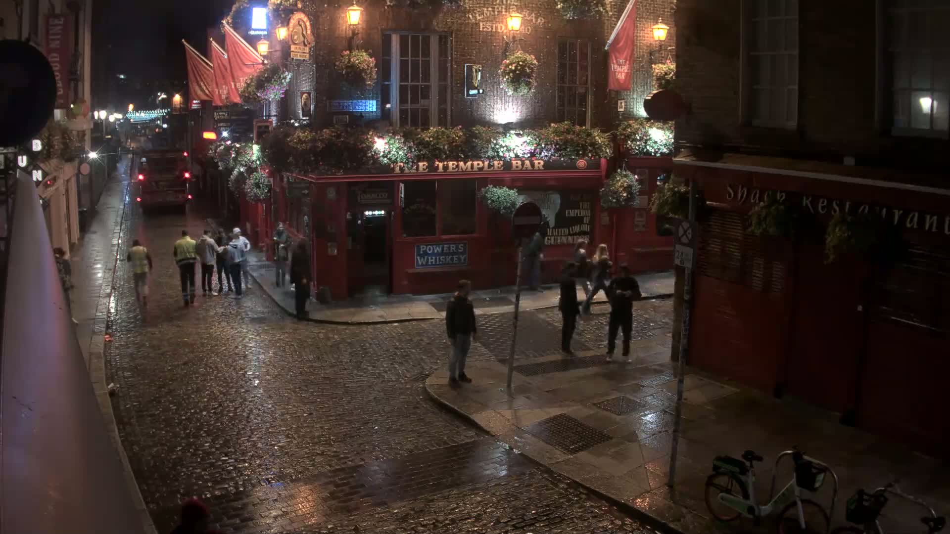 A rain-slicked cobblestone street at night shows several people walking near a red brick pub and restaurant.