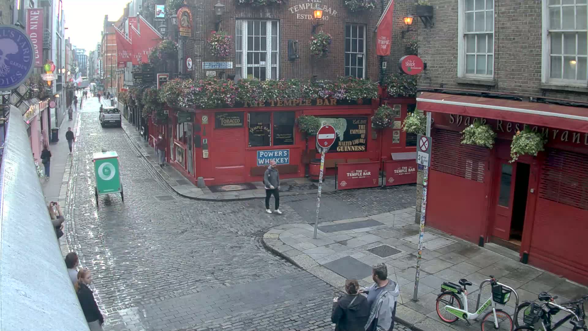 A wet cobblestone street in a city, with red brick buildings, pedestrians, and a delivery cart, under an overcast sky.