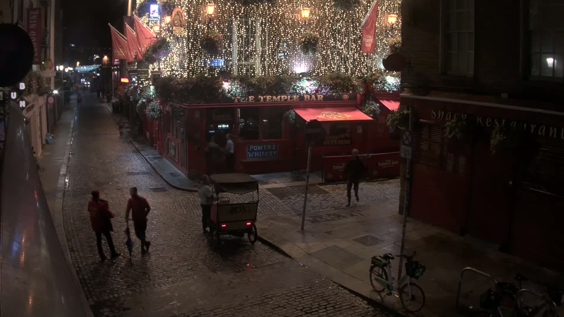 A wet, cobblestone street at night is lined with buildings, one of which is brightly lit and decorated with string lights, with several people walking and a rickshaw parked nearby.