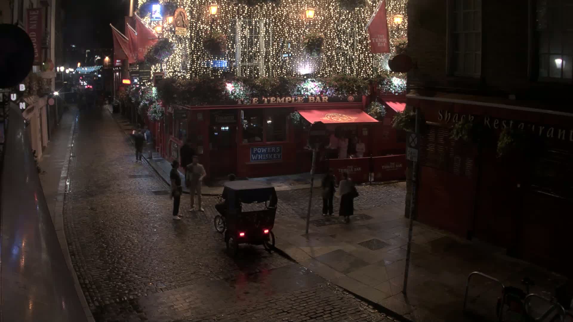 A rain-slicked cobblestone street at night, lined with buildings, one of which is brightly lit and adorned with string lights, features a rickshaw and several pedestrians.