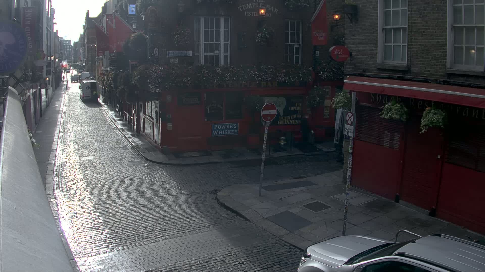 A cobblestone street, wet from recent rain, runs between historic buildings, one of which is a pub, and a silver SUV is parked at the bottom of the street.