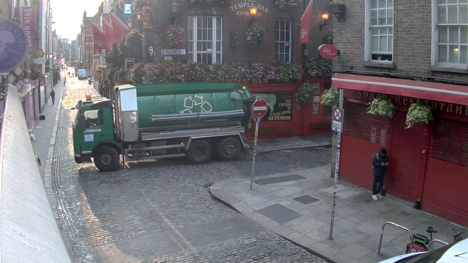 A green recycling truck is parked on a cobblestone street in front of a red building on a partly sunny day.