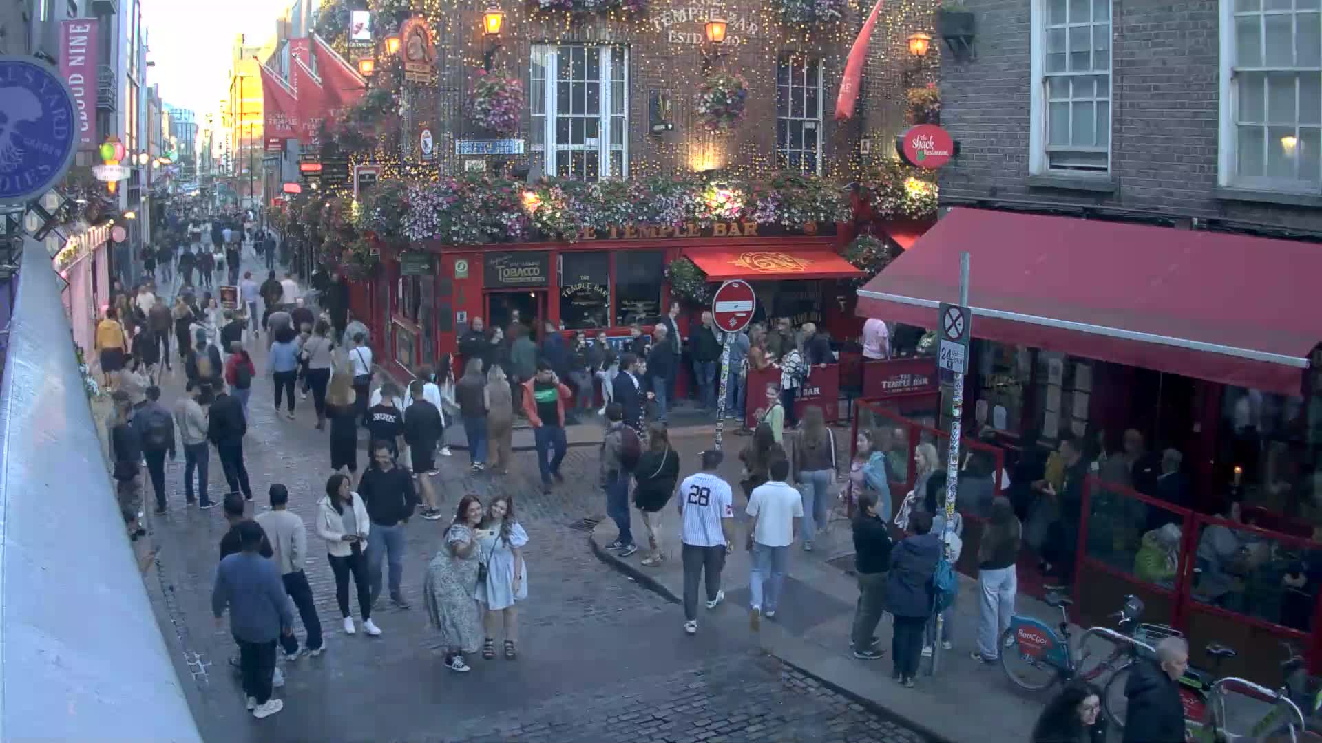 A crowded cobblestone street at dusk, with many people walking past a brightly lit red pub, under a mostly clear sky.