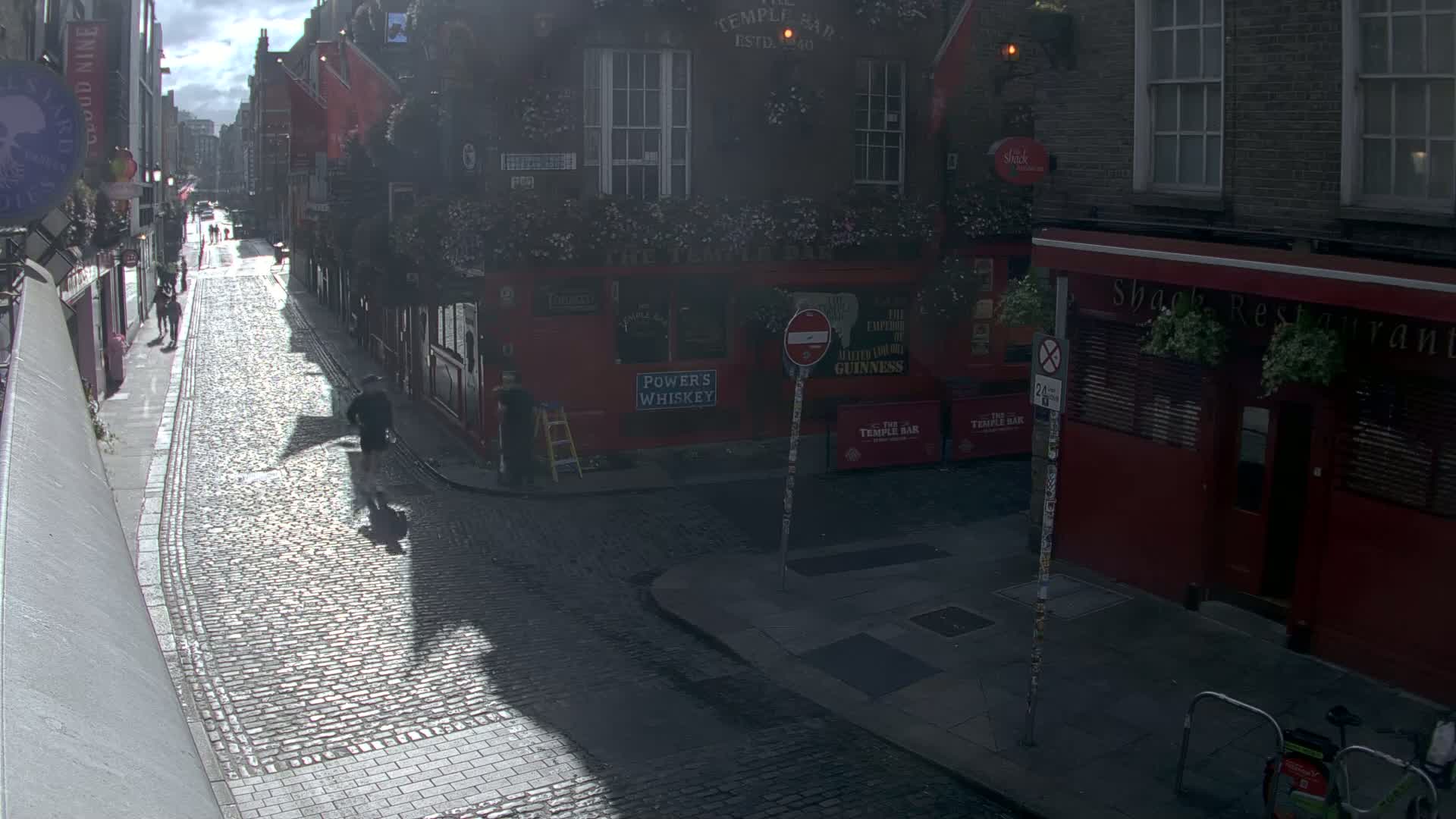 A cobblestone street lined with red brick buildings, under a partly sunny sky, features several pedestrians and a person working near a ladder.