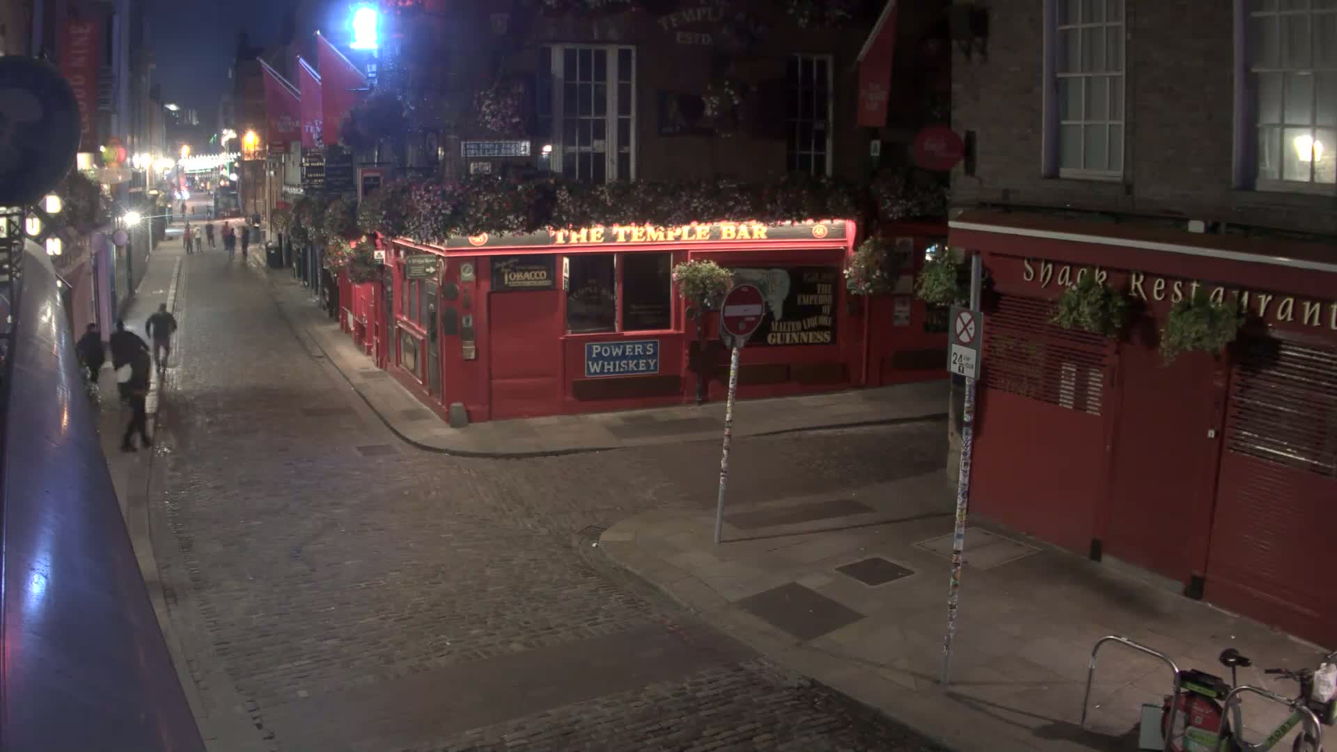 A cobblestone street at night, with several people walking past red brick buildings, one of which is a bar.