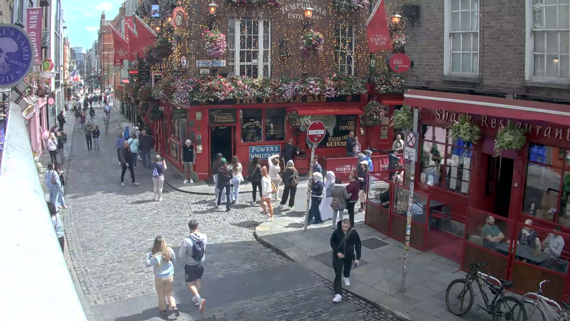 A cobblestone street, bustling with pedestrians, features red brick buildings, including a prominent pub adorned with hanging flowers under a sunny sky.