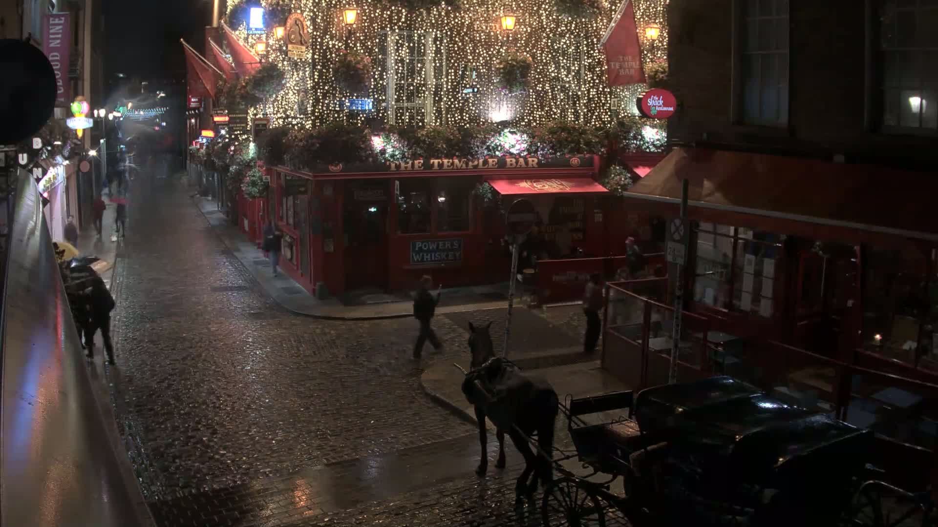 A rain-slicked cobblestone street at night, lined with buildings, one of which is brightly lit and decorated with string lights, with a horse-drawn carriage and pedestrians visible.