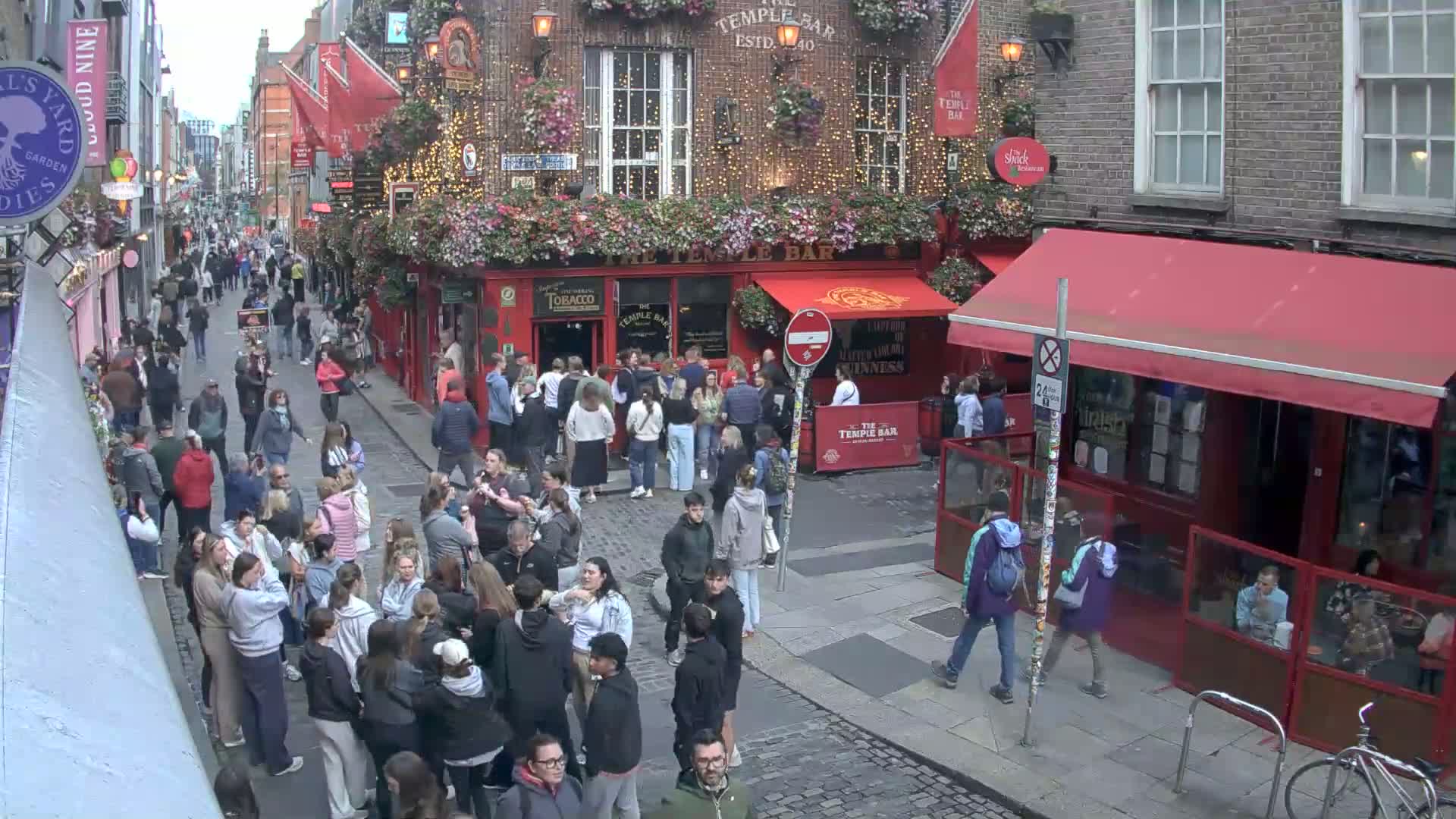 A crowded cobblestone street in daylight, with people walking past and standing outside of a red-fronted pub.