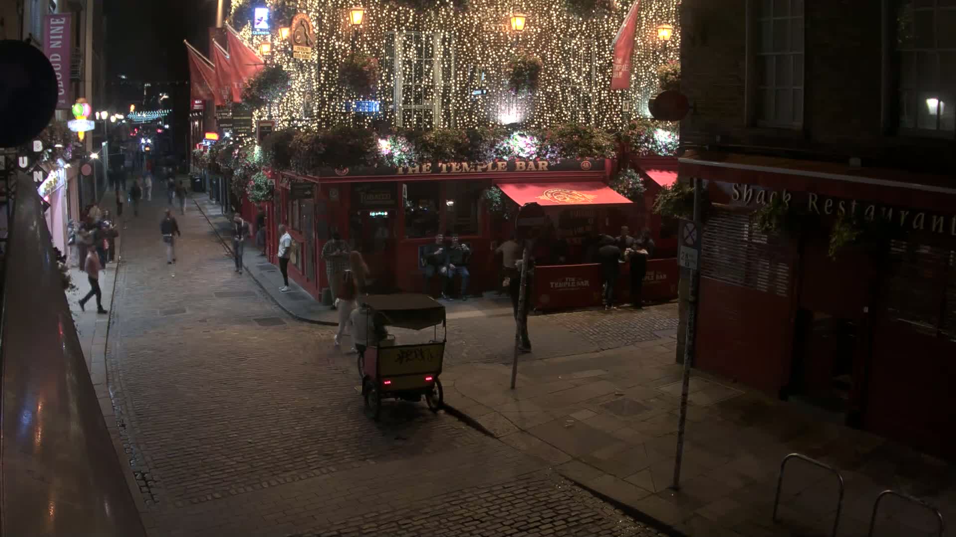 A cobblestone street at night, lined with illuminated buildings, including a brightly lit pub adorned with string lights, and several pedestrians are visible.