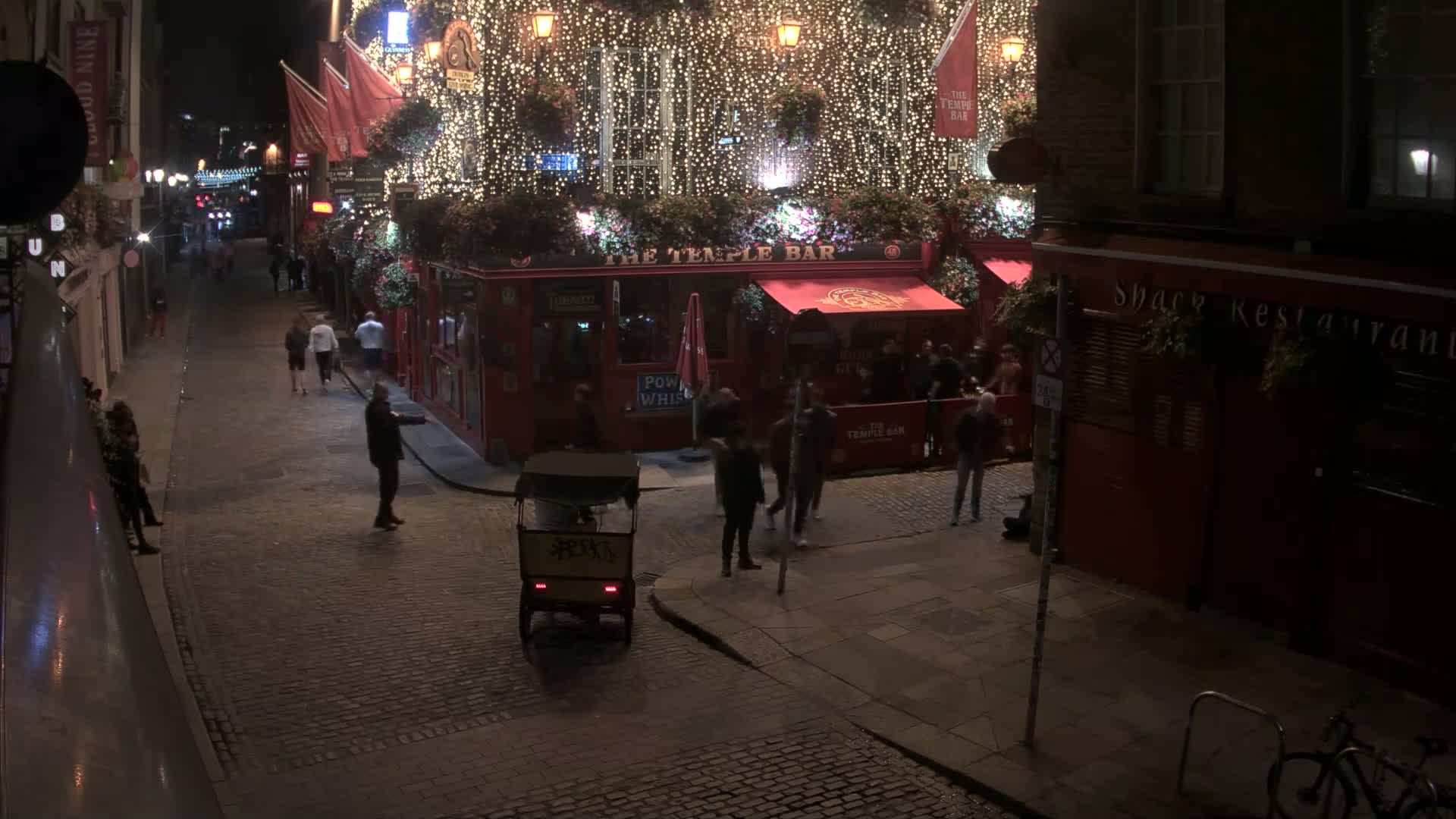 At night, people stroll along a cobblestone street past a brightly lit pub decorated with string lights, and a rickshaw sits nearby.