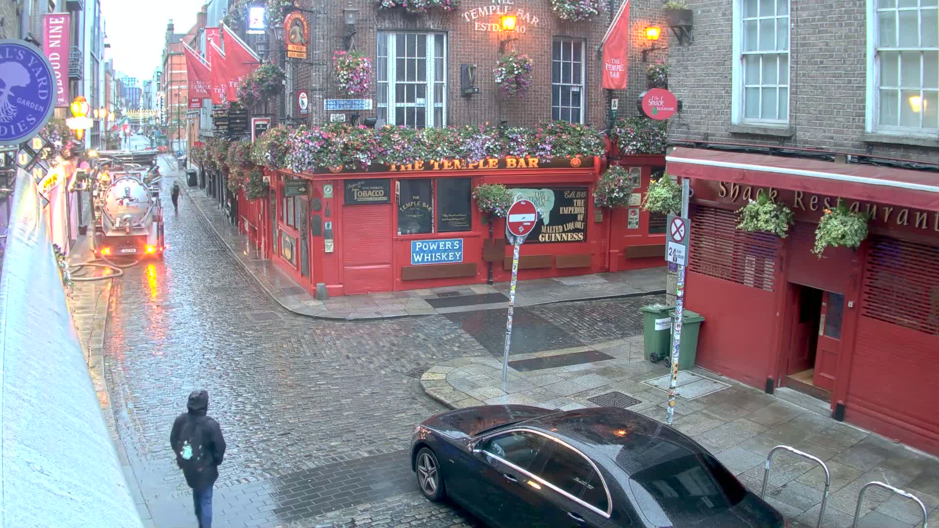 A wet, cobblestone street scene shows a red brick building with flower boxes, a parked black car, and a person walking past a street cleaning truck in the rain.