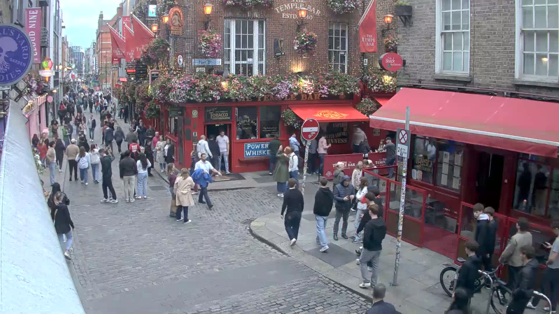 A cobblestone street filled with people, some near a red-fronted pub with hanging flower baskets, under an overcast sky.