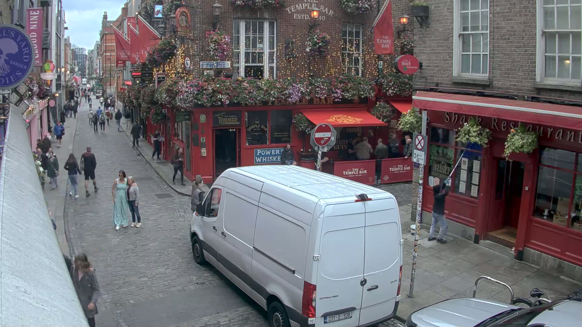 A cobblestone street scene shows a white van driving past a red-brick building with flower boxes, pedestrians walking, and overcast sky conditions.