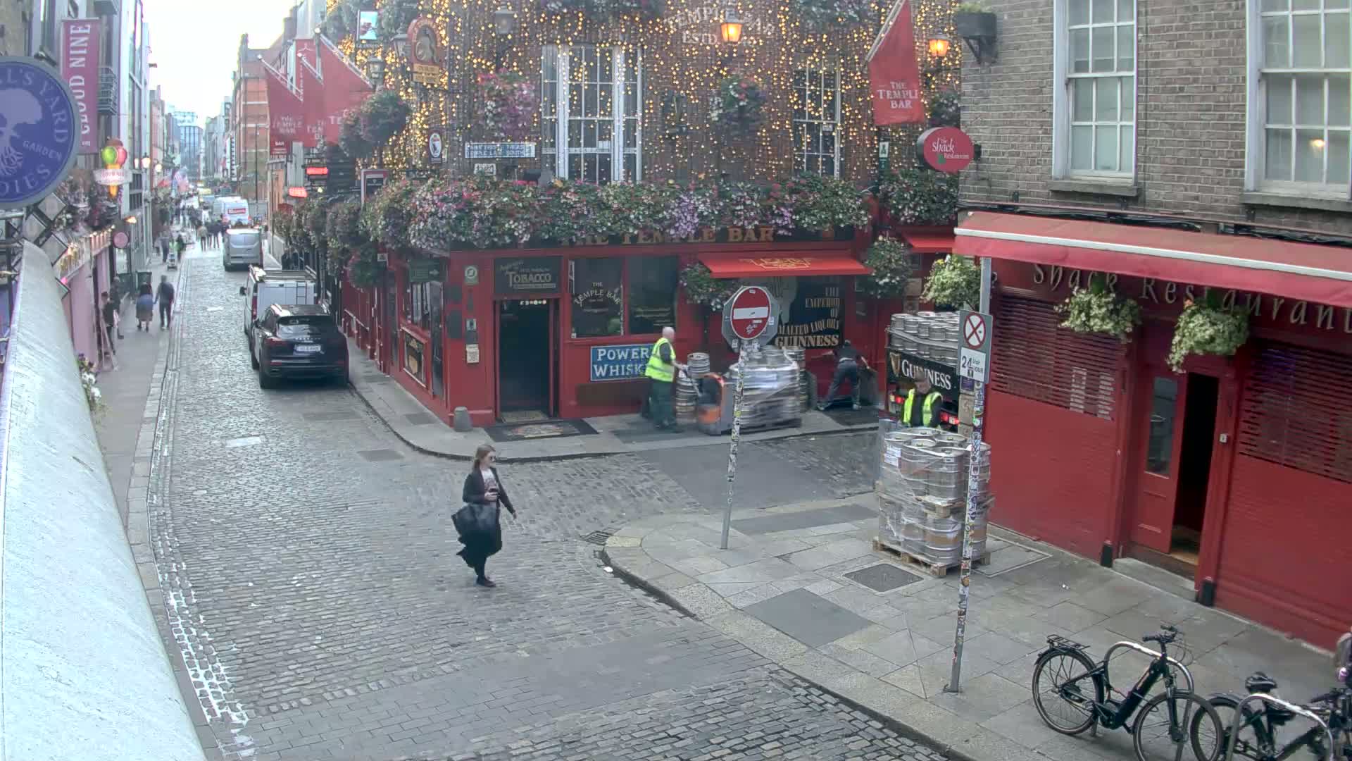 A cobblestone street, possibly in a European city, shows overcast weather conditions, with pedestrians walking past red brick buildings, one of which is a pub with outdoor seating and hanging plants, where workers are loading kegs.