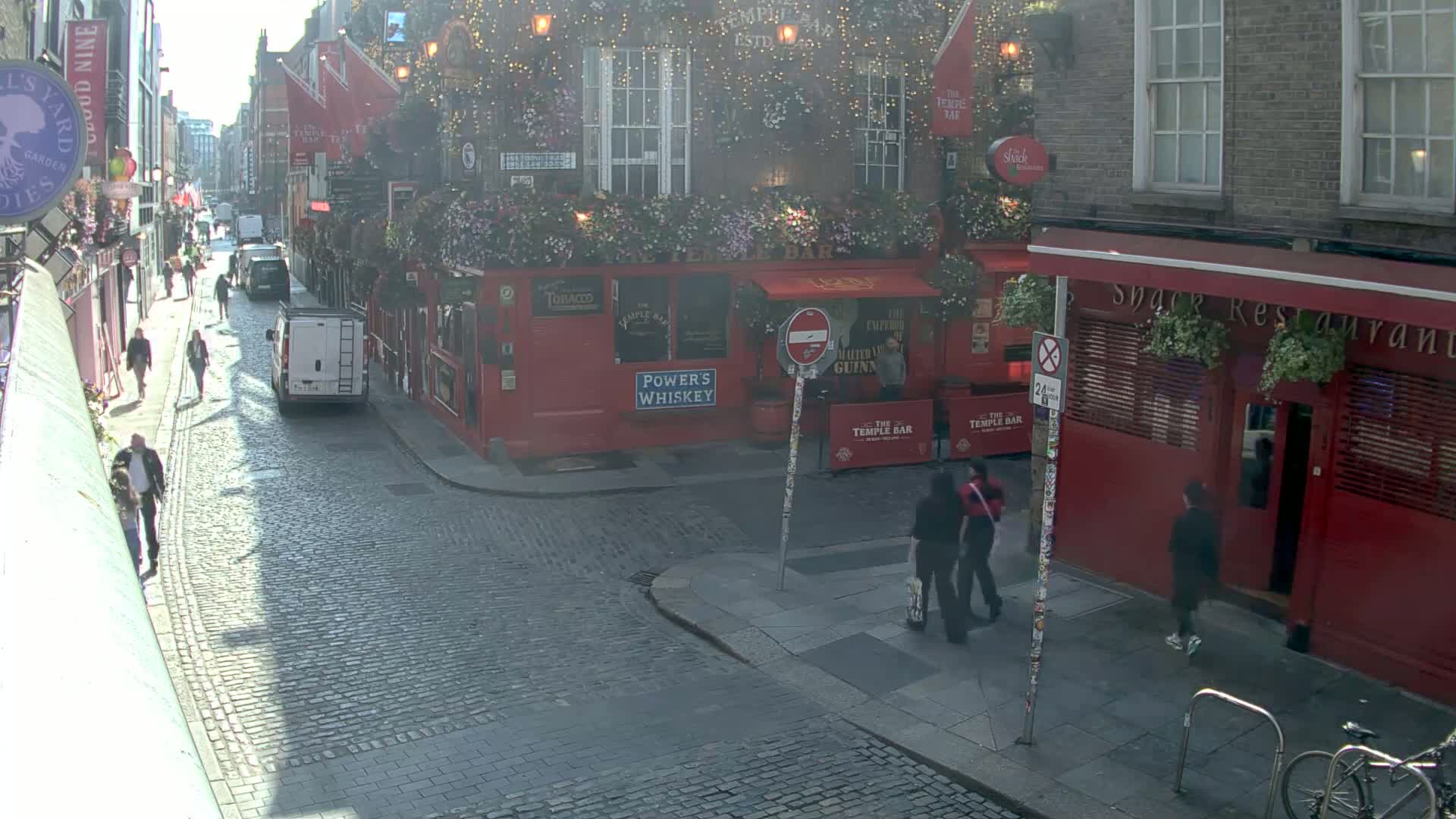 A cobblestone street scene on a sunny day shows pedestrians walking past red brick buildings with pubs and restaurants.