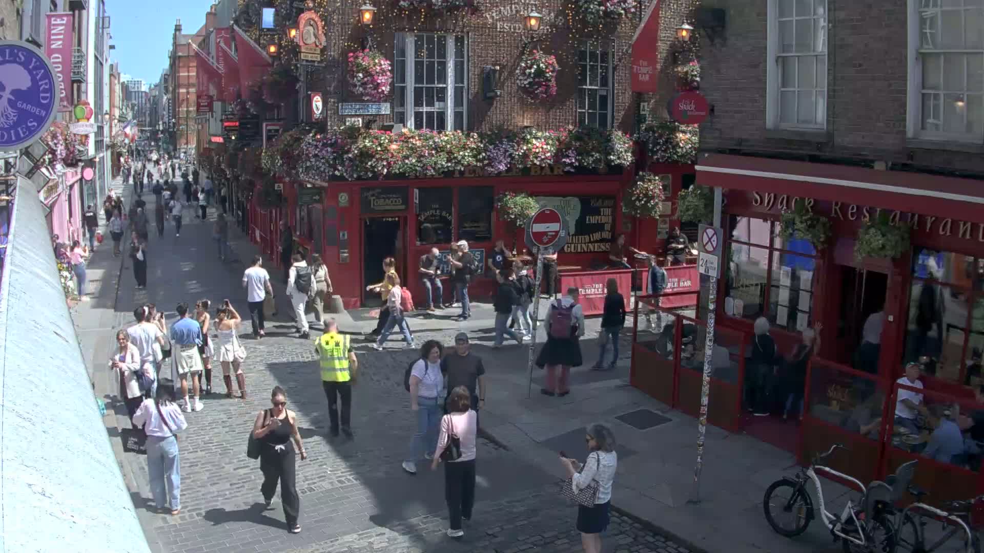 A sunny cobblestone street is filled with pedestrians walking past shops and pubs, one of which is the Temple Bar.