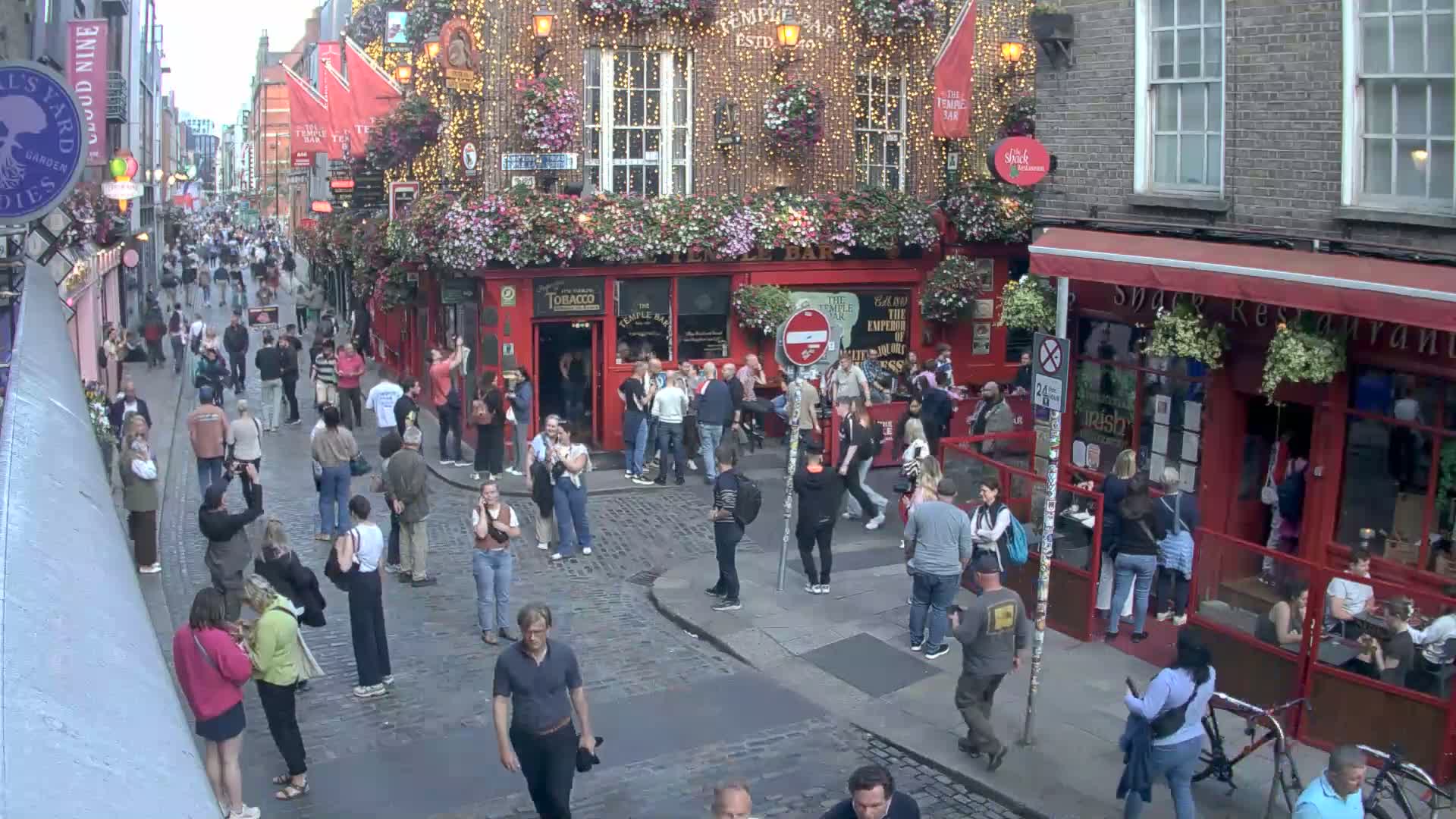 A bustling cobblestone street, filled with pedestrians, features a prominent red-brick building adorned with flowers and string lights under a twilight sky.