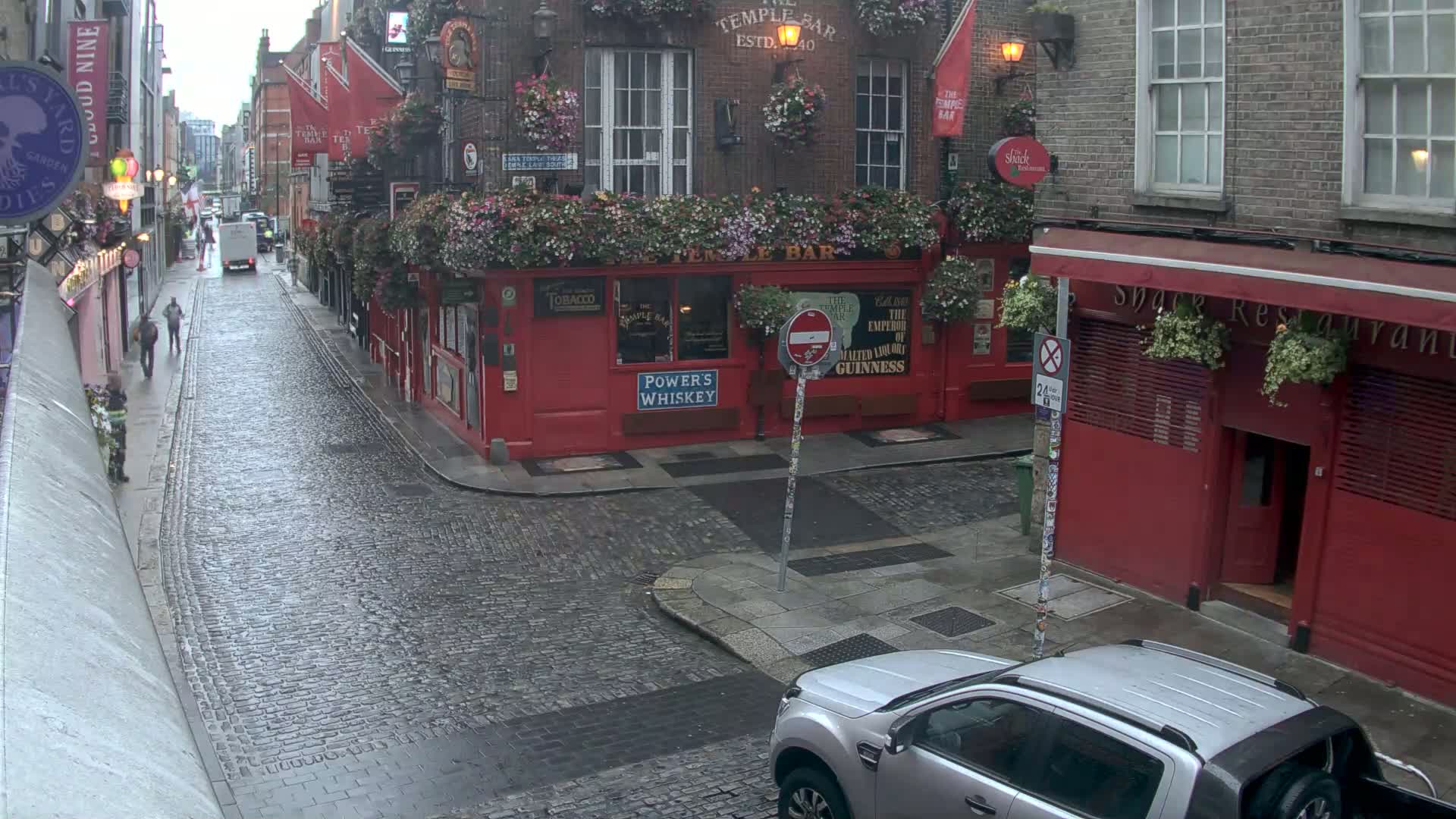A rain-slicked cobblestone street in a city, lined with red brick buildings, some with flower boxes and signage.
