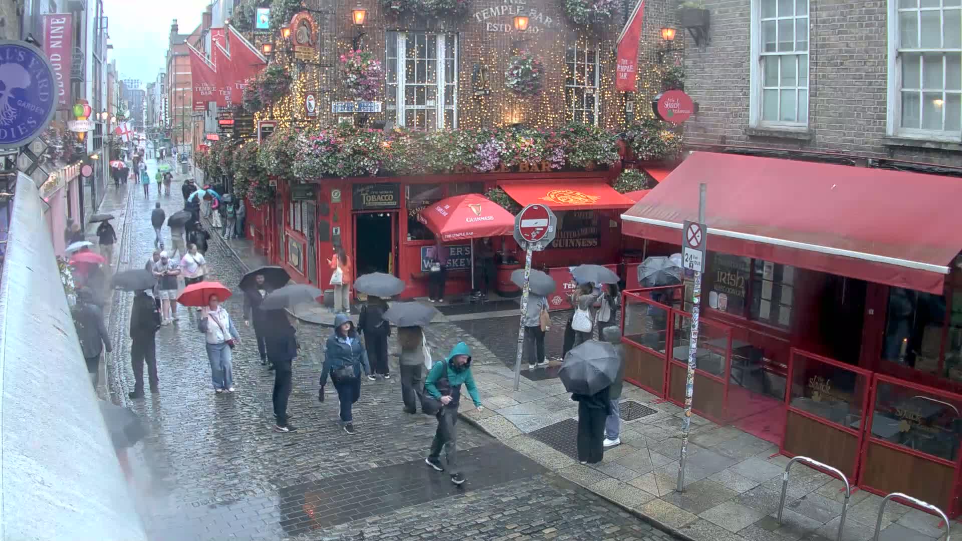 A rainy cobblestone street scene shows numerous people with umbrellas walking past red-awninged pubs and shops.