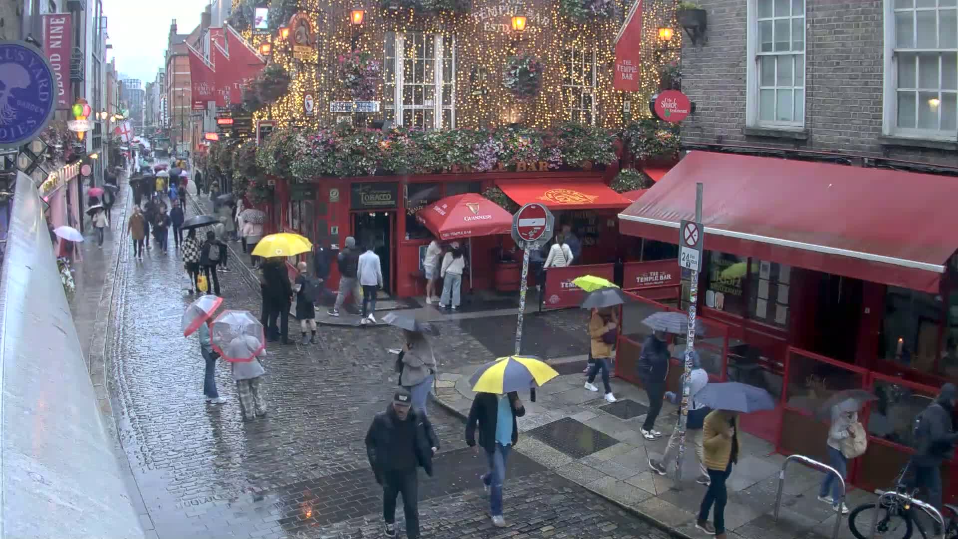 A rain-soaked cobblestone street is filled with people carrying umbrellas, walking past pubs and shops in a city setting.