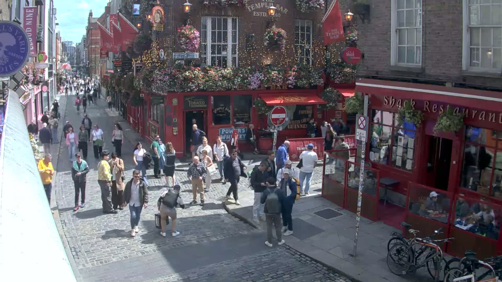 A cobblestone street scene on a sunny day shows many people walking past pubs and restaurants with hanging flower baskets.