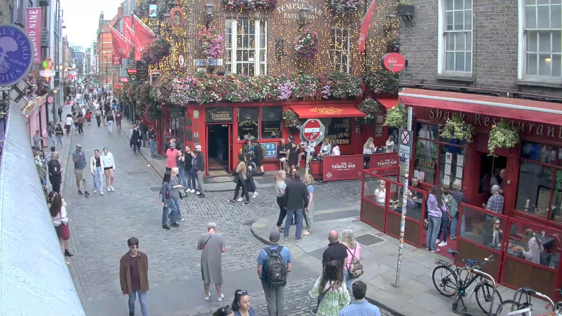 A cobblestone street scene shows many people walking past numerous pubs and restaurants on a sunny day.