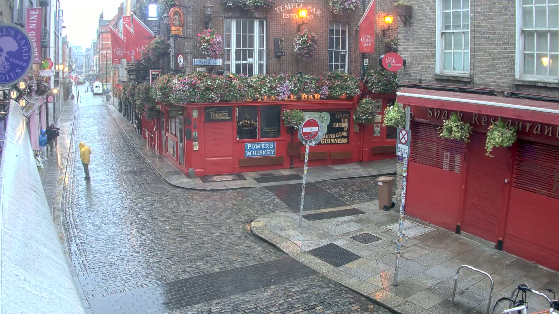 A cobblestone street scene shows many people walking past numerous pubs and restaurants on a sunny day.