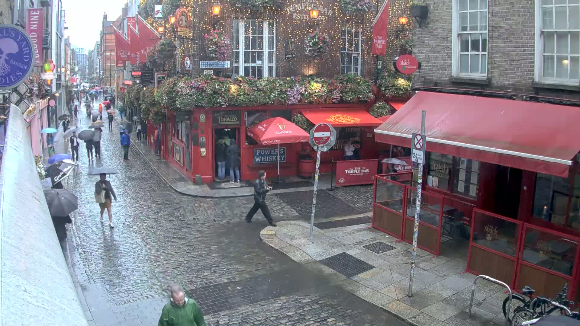 A rain-slicked cobblestone street scene shows pedestrians with umbrellas walking past red-brick buildings and pubs decorated with flowers and string lights.