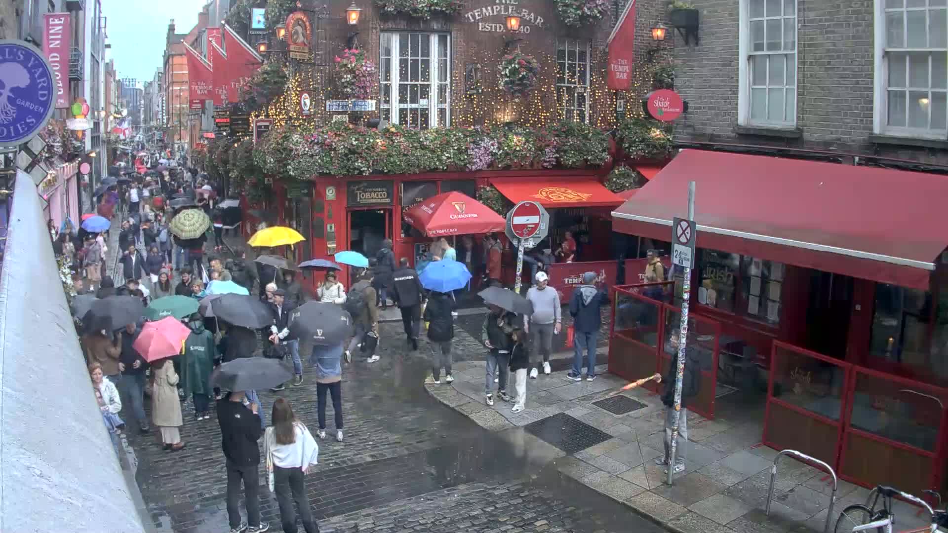 A crowded, cobblestone street with many people carrying umbrellas under a rainy sky, in front of a red-awninged pub.
