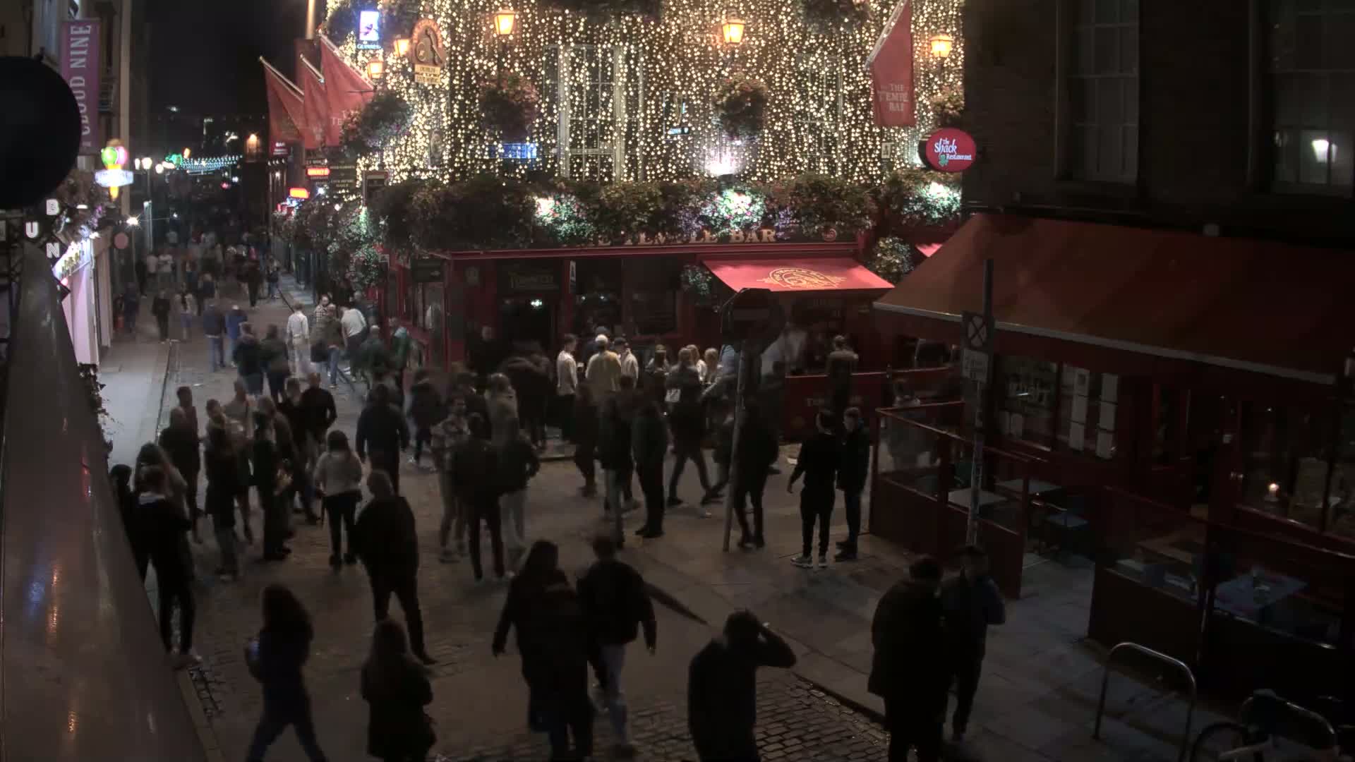 A crowded, cobblestone street with many people carrying umbrellas under a rainy sky, in front of a red-awninged pub.