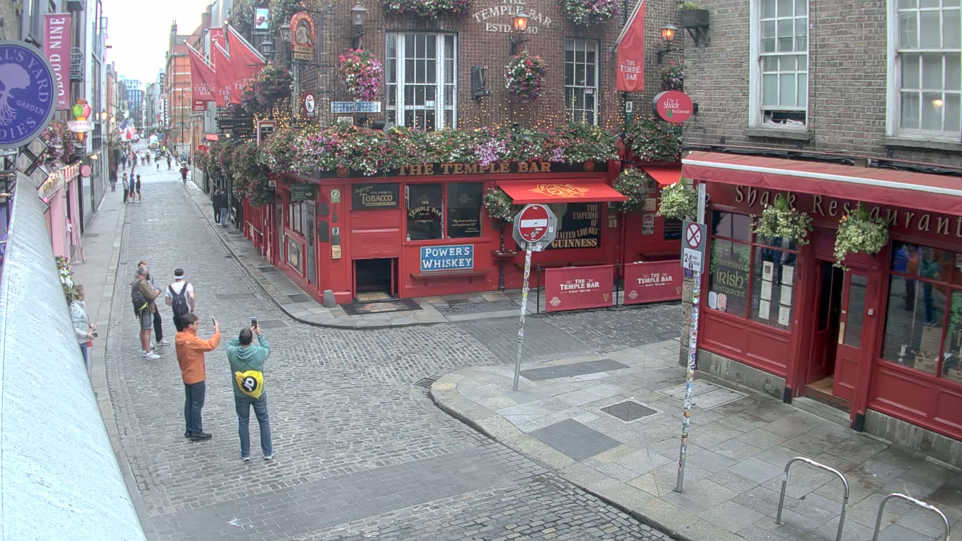 A cobblestone street in overcast weather features several people taking photos near red brick buildings with flower boxes and pub signs.