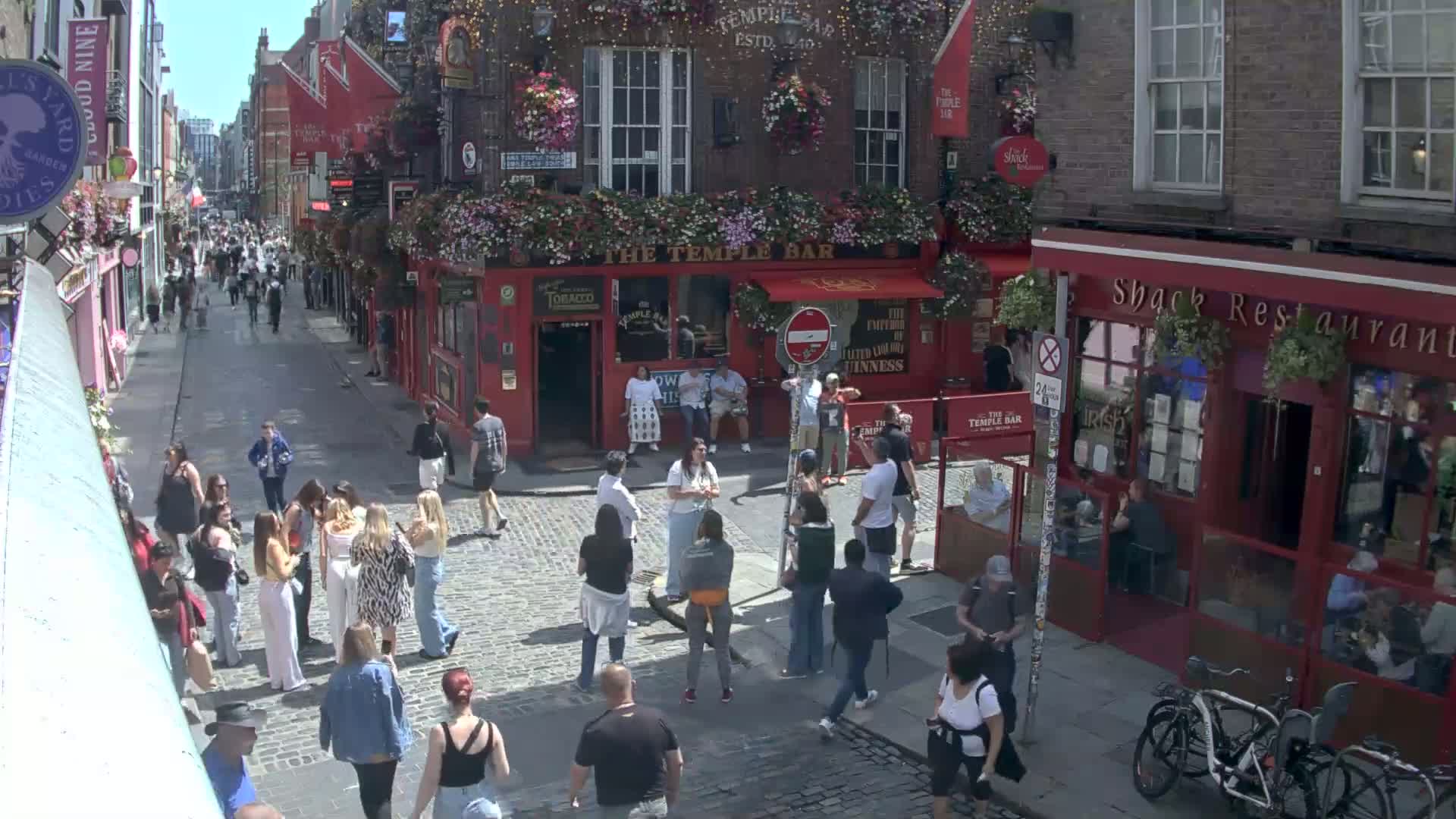 A sunny cobblestone street in a city, filled with pedestrians, and lined with red brick buildings, one of which is a pub.