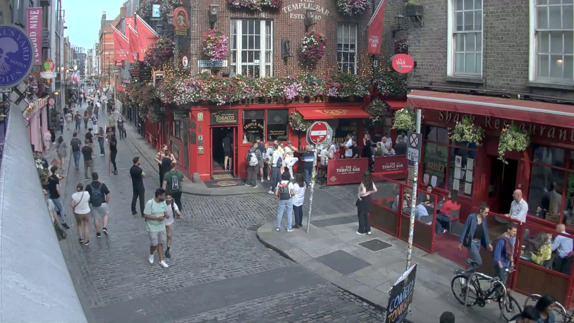 A cobblestone street, bustling with pedestrians, features a red-brick pub with outdoor seating and hanging flower baskets under a bright, sunny sky.