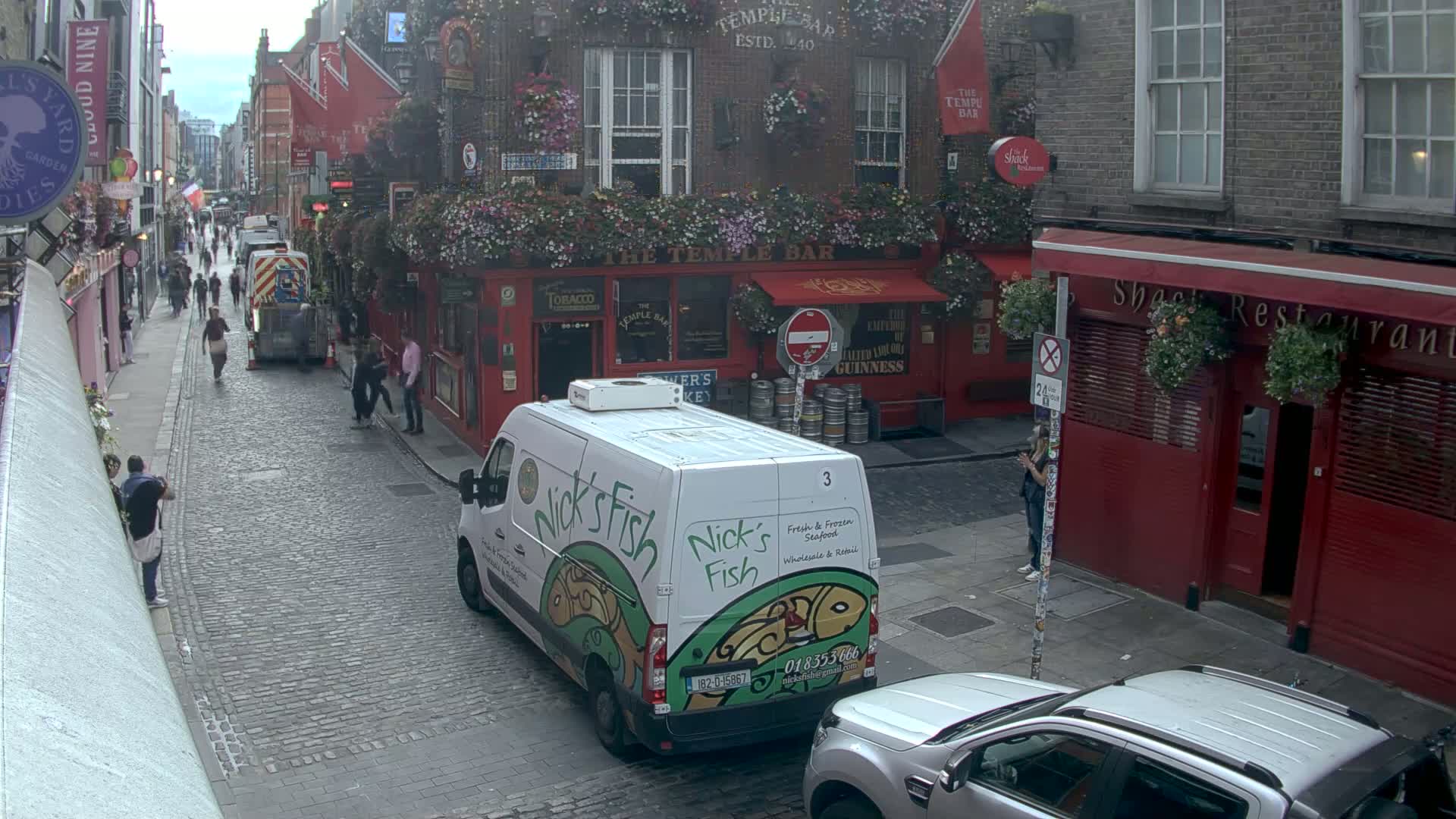 A cobblestone street scene on an overcast day shows a white delivery van parked near a red brick building with hanging flower baskets, and pedestrians walking past.