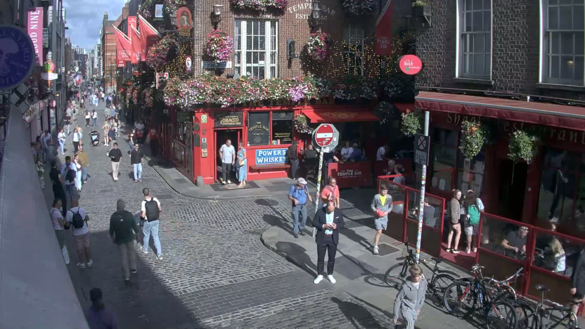 A sunny cobblestone street in a city, filled with pedestrians walking past numerous buildings, including a red pub with flower boxes, and several bicycles parked along the side.