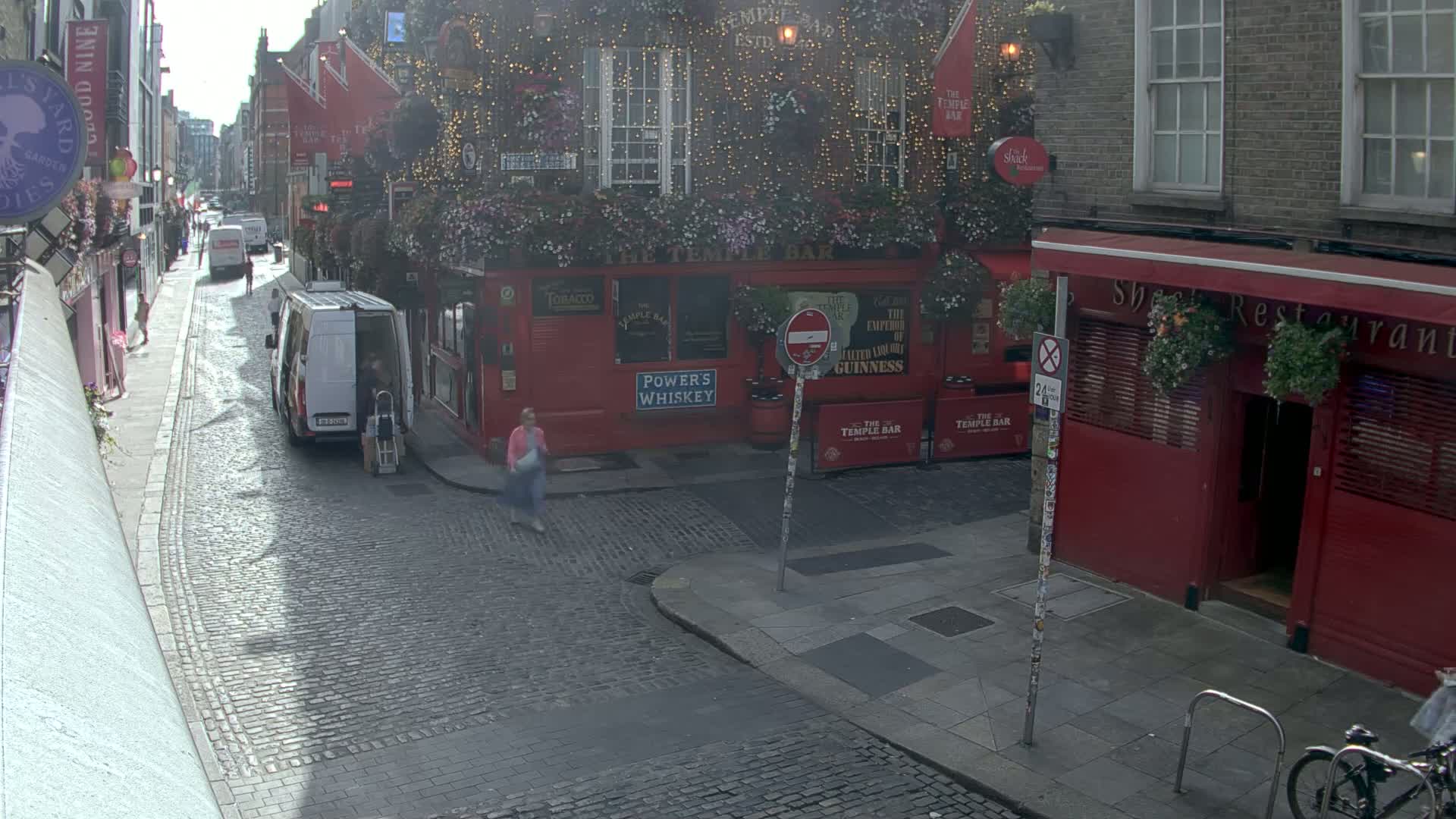 A cobblestone street scene on a sunny day shows a red building with hanging plants and string lights, a van parked nearby, and a few pedestrians walking.