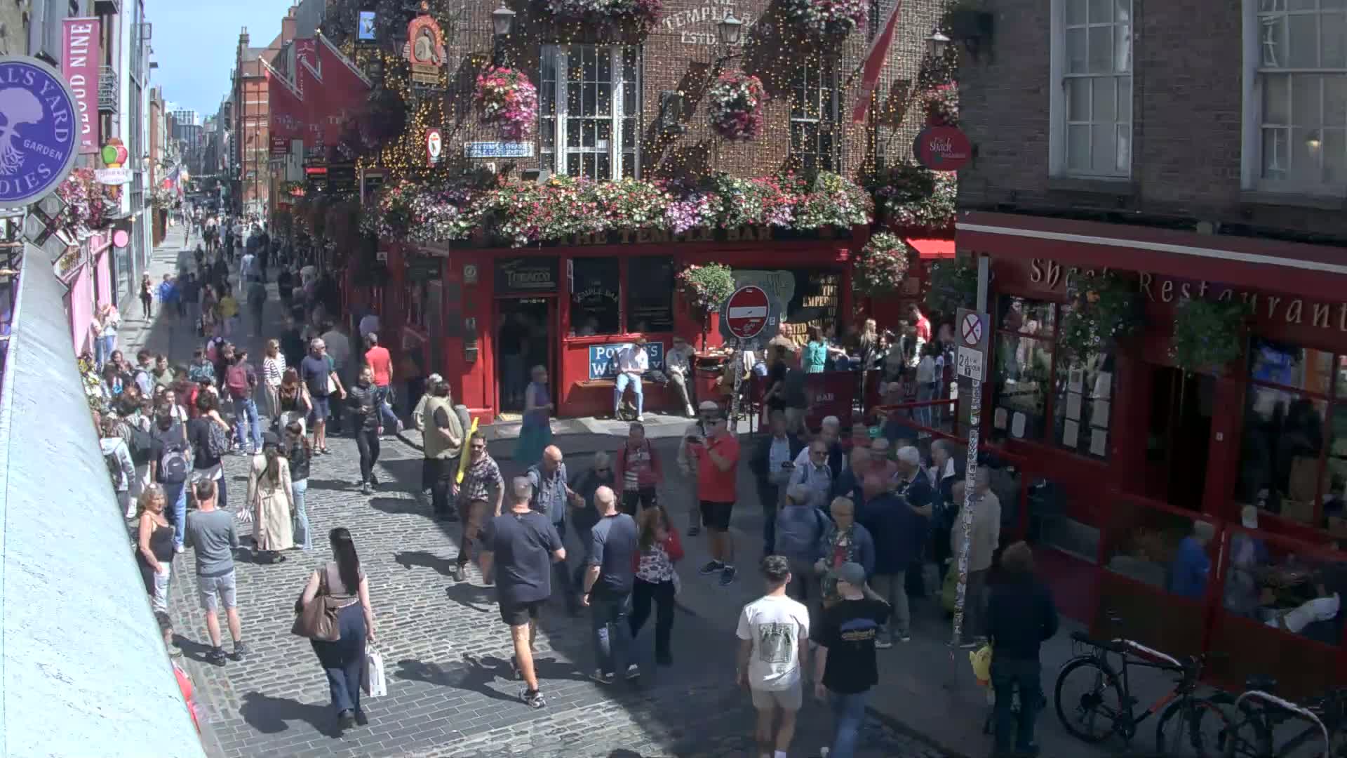 A crowded cobblestone street with numerous pedestrians, flanked by red brick buildings, under sunny conditions.