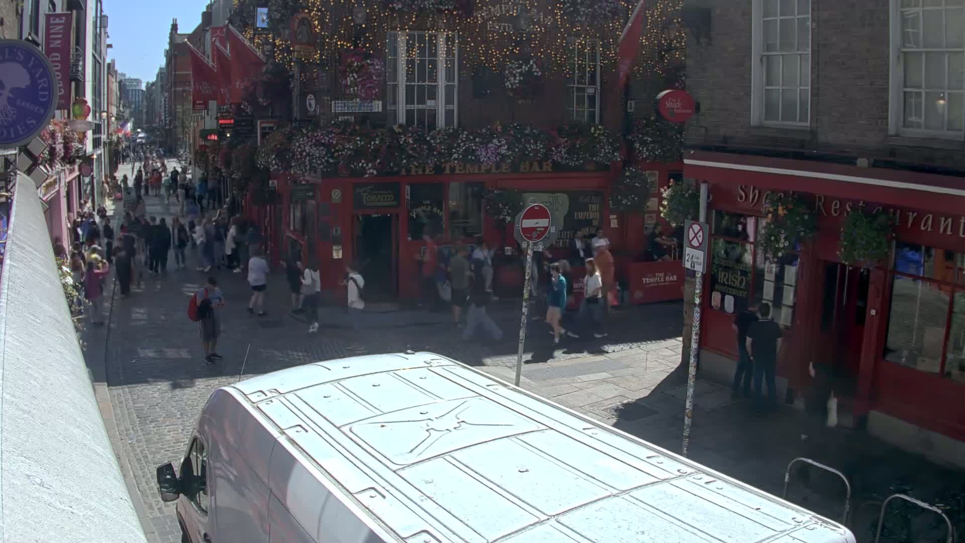 A crowded cobblestone street on a sunny day, with a white van in the foreground and numerous people walking past red brick buildings and pubs.