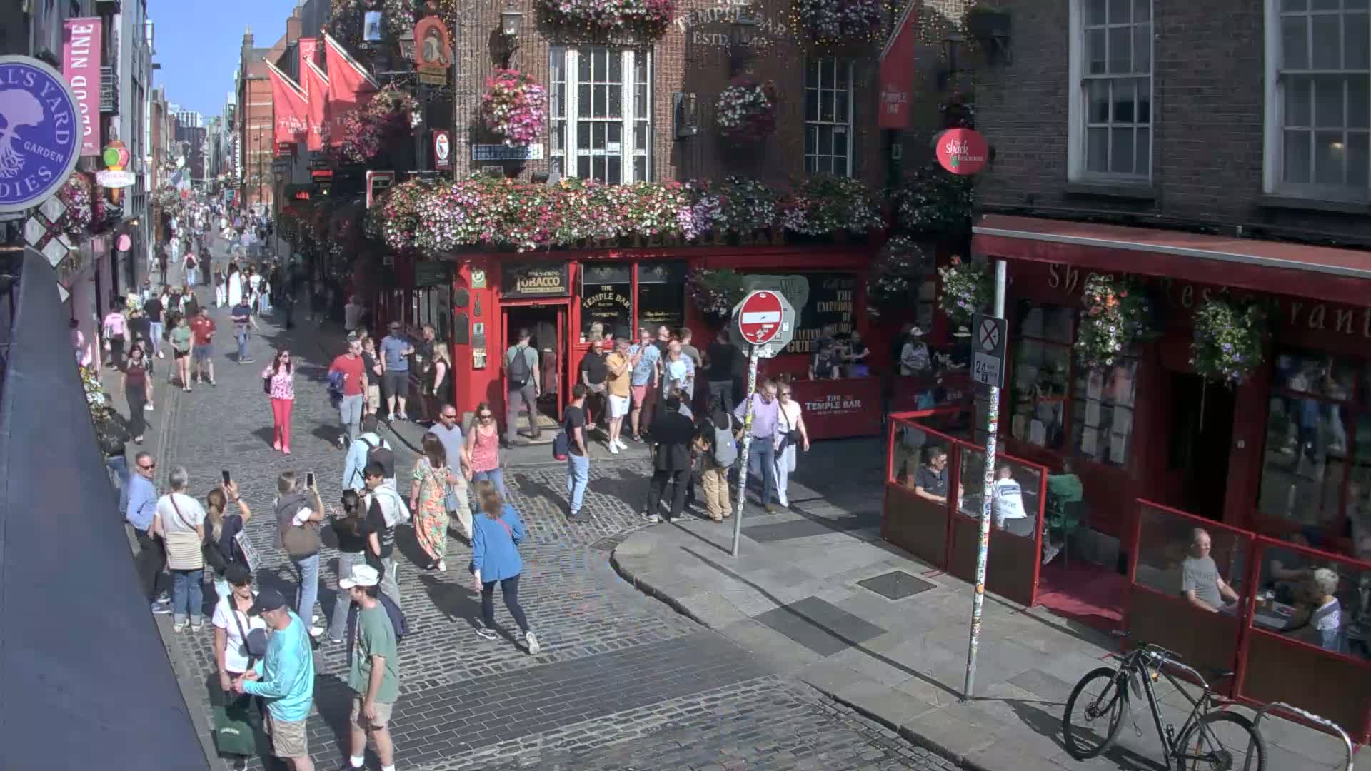 A sunny day on a cobblestone street shows many pedestrians walking past several red brick buildings with flower boxes and outdoor seating areas.
