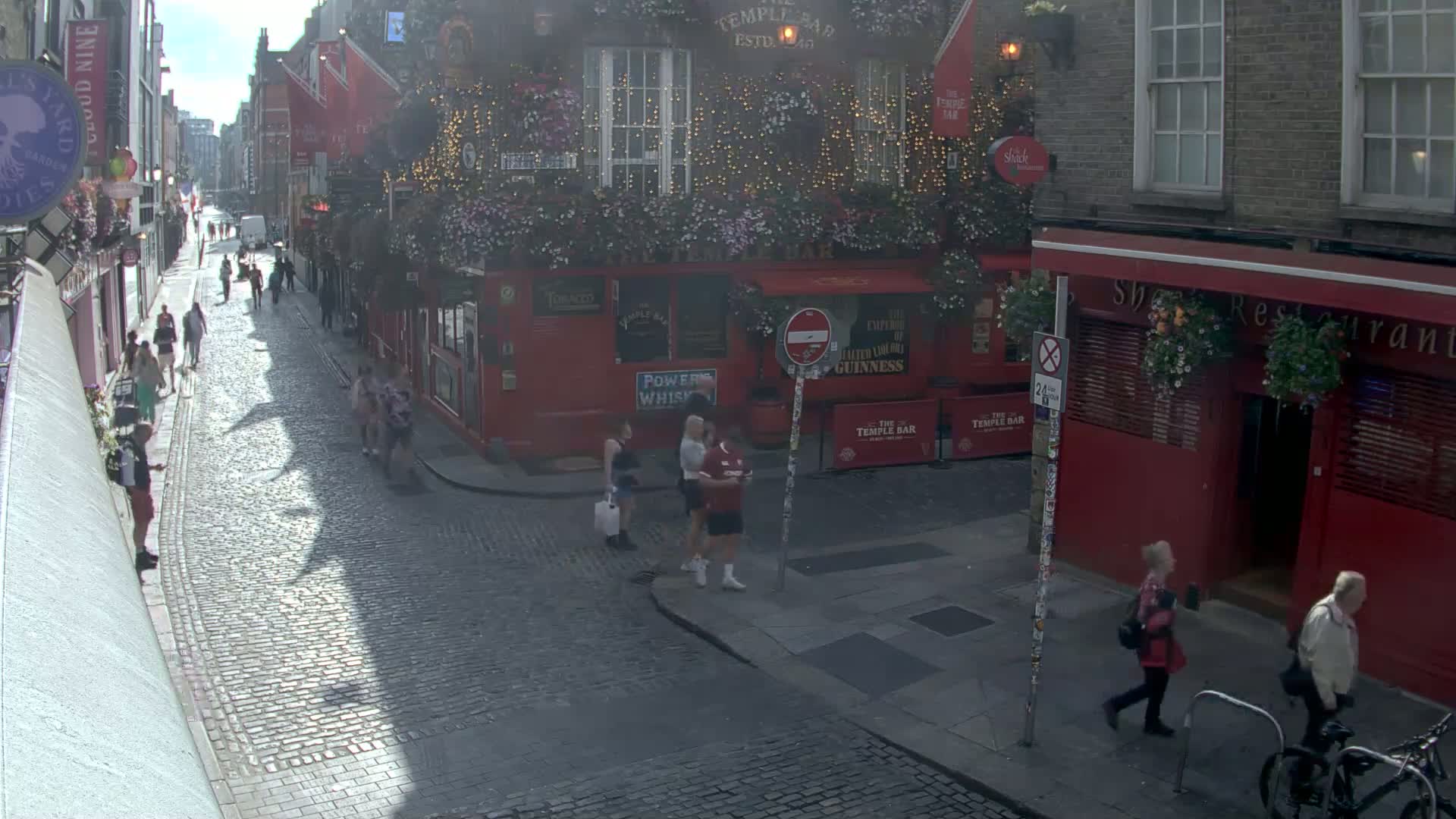A sunny cobblestone street with pedestrians and red brick buildings, including a pub decorated with flowers and string lights.