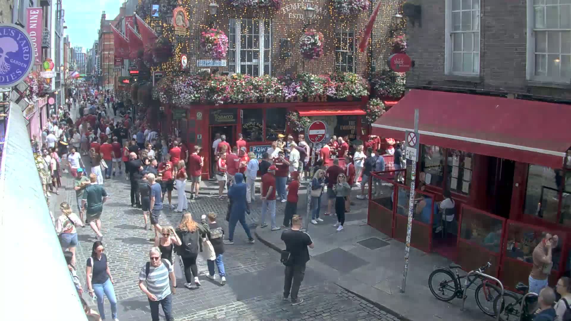 A crowded cobblestone street on a sunny day, filled with people, many wearing red, near a red-awninged pub adorned with hanging flowers.