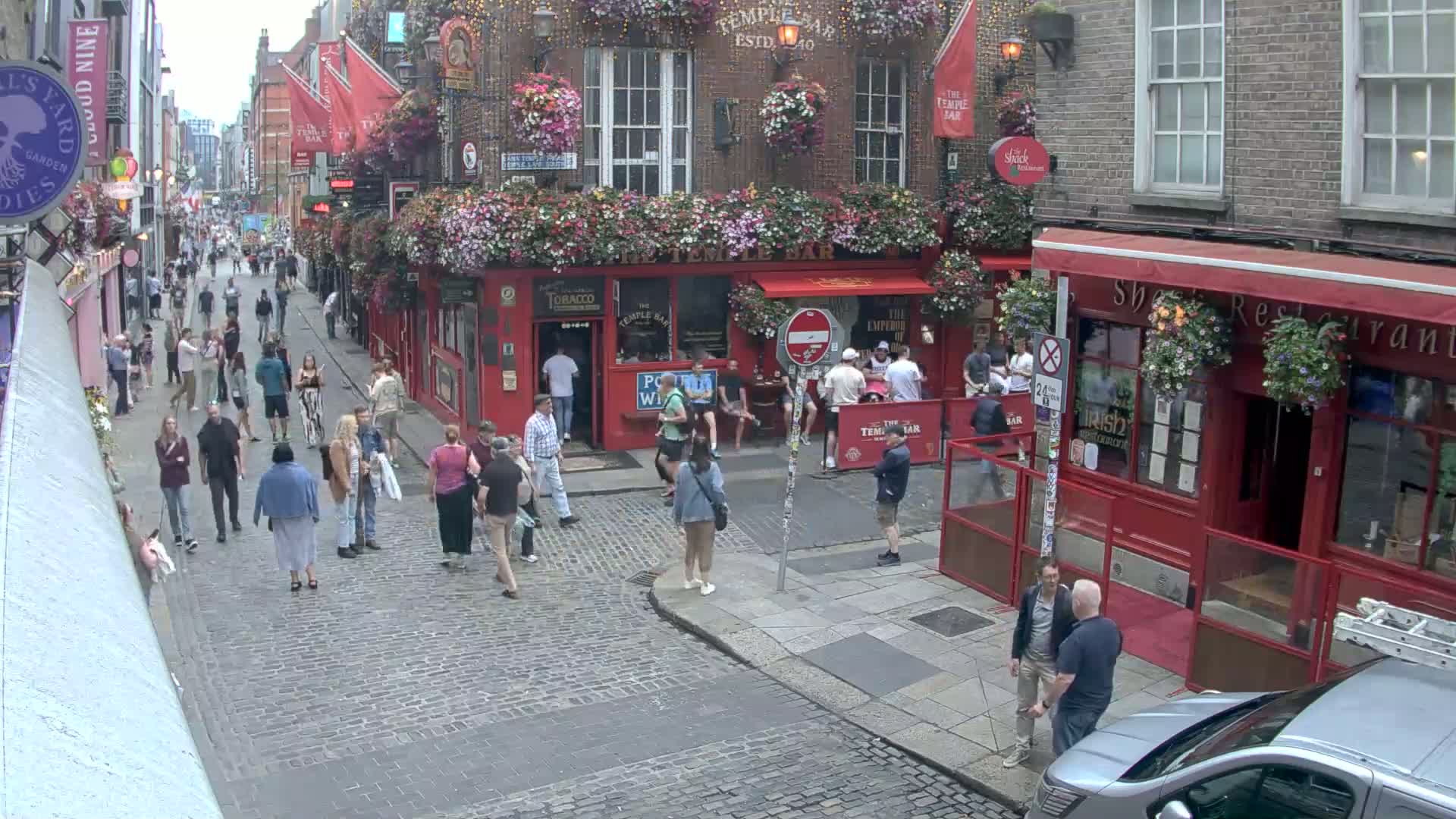 A cobblestone street lined with red-brick buildings, many with flower boxes, is bustling with pedestrians on a partly cloudy day.