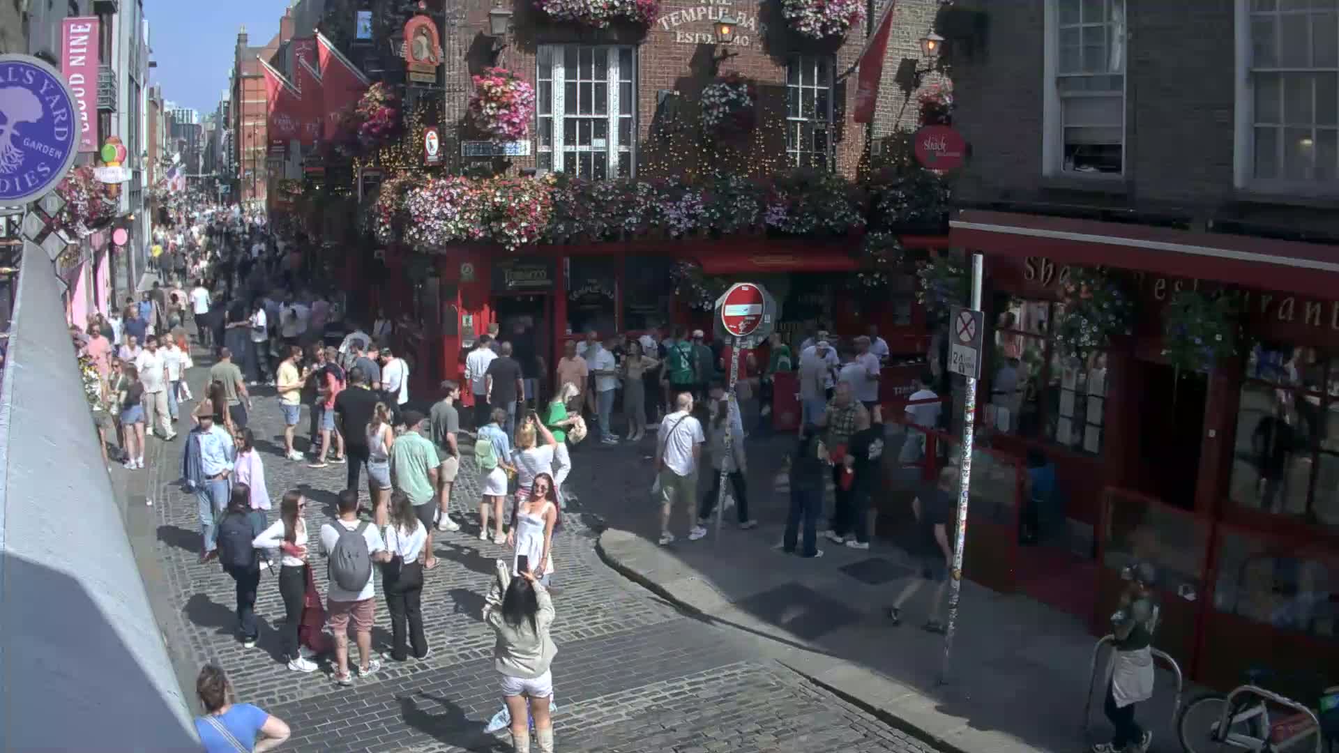 A crowded cobblestone street scene on a sunny day features numerous pedestrians walking past buildings with hanging flower baskets.