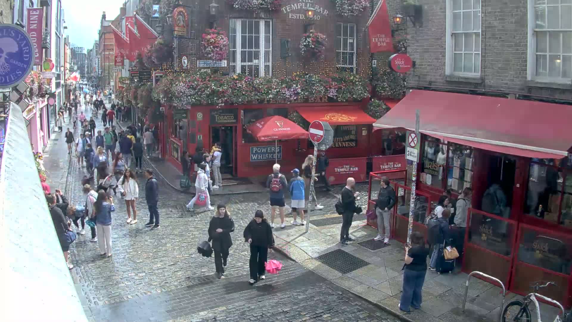 A rain-slicked cobblestone street, crowded with pedestrians, runs past several red-brick buildings with awnings and outdoor seating.