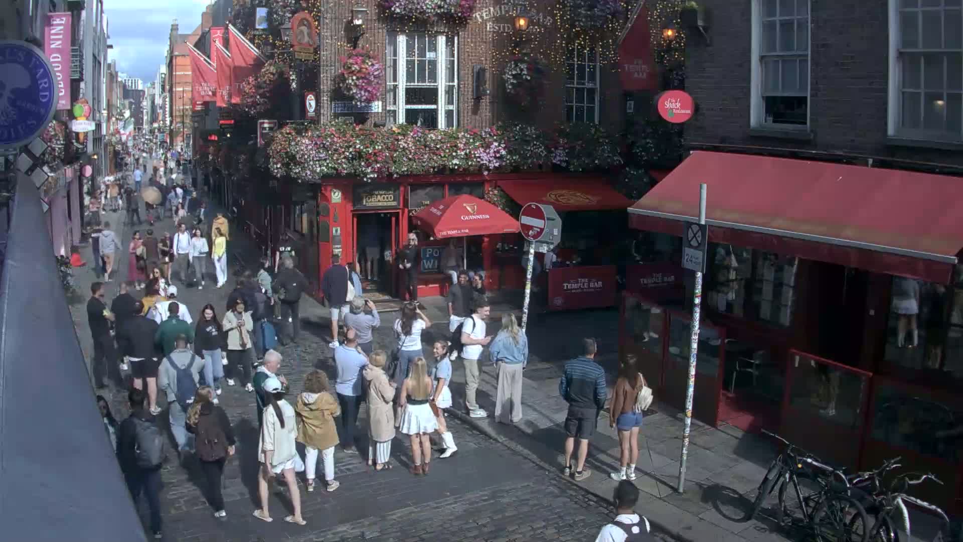 A sunny day on a cobblestone street with many people walking past several red brick buildings, some with awnings and flower boxes.