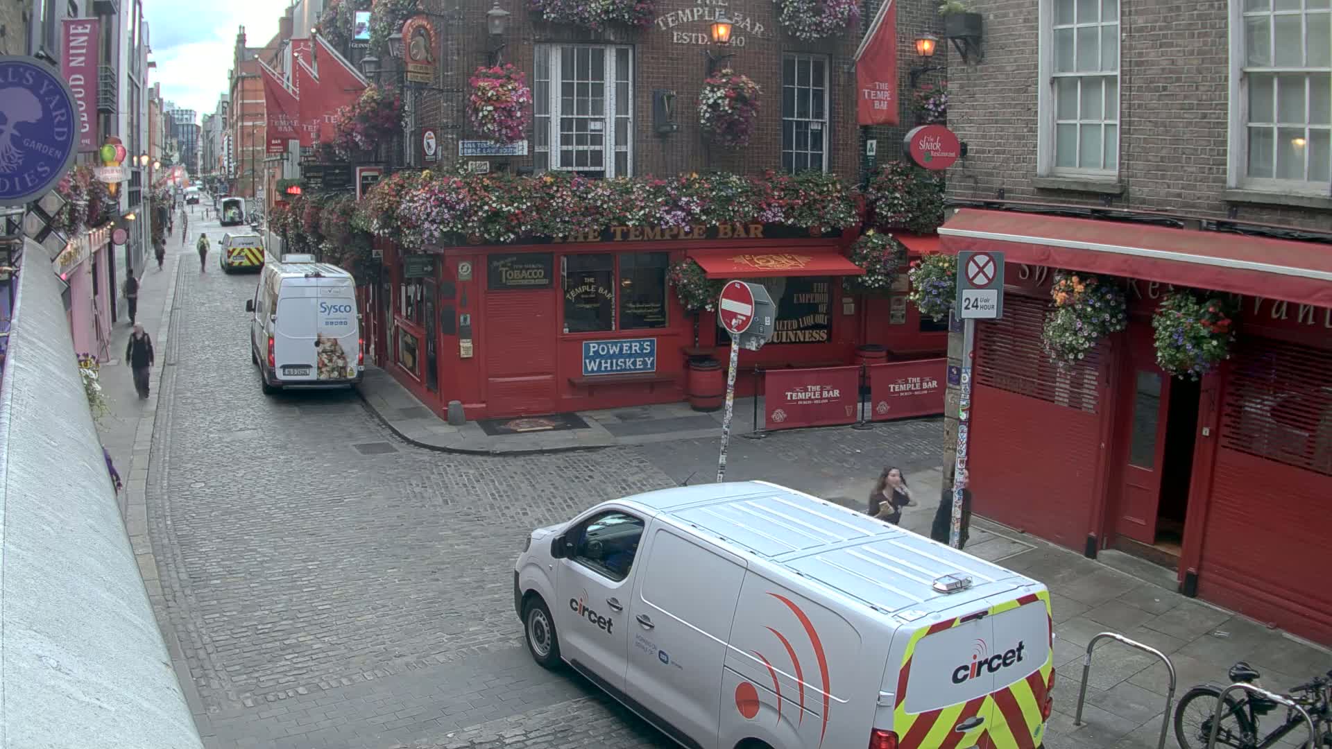 A cobblestone street with red brick buildings, flower boxes, and several vans, on an overcast day.