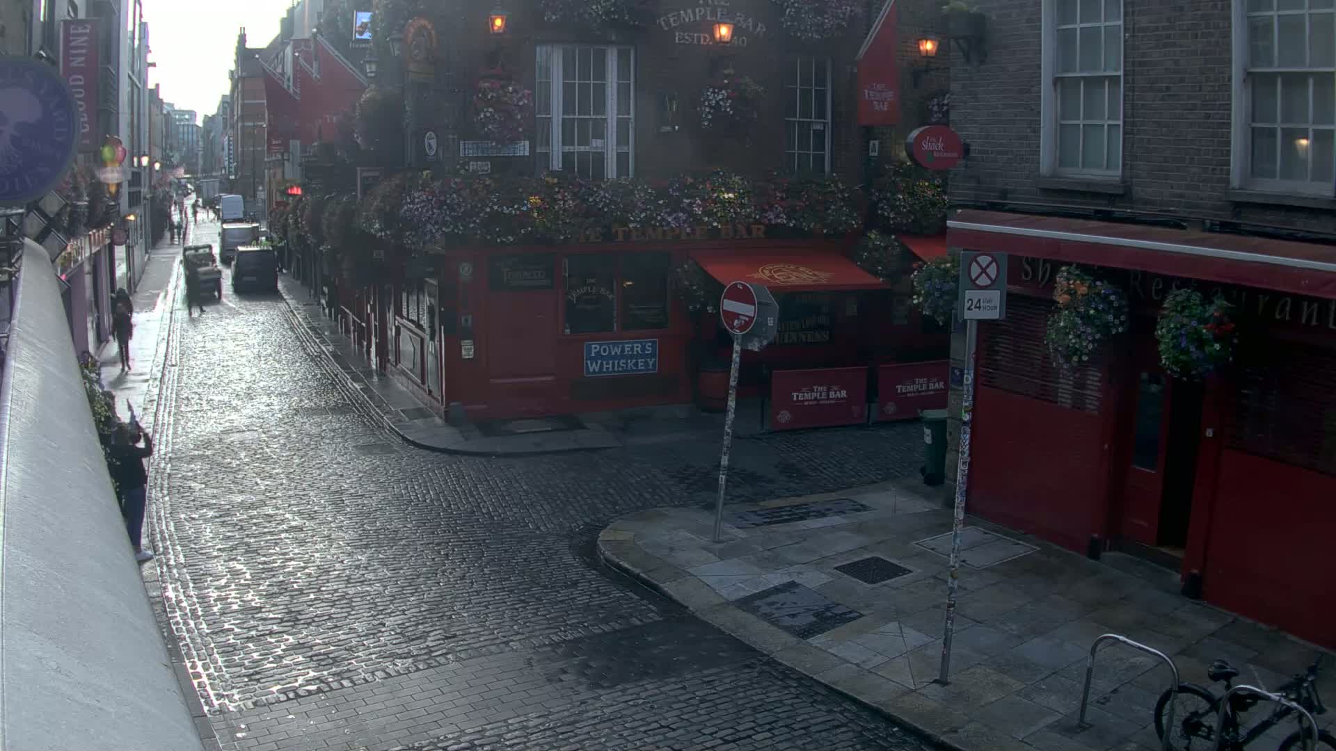 A wet cobblestone street in a city with red brick buildings and hanging flower baskets, showcasing a pub called "The Temple Bar".