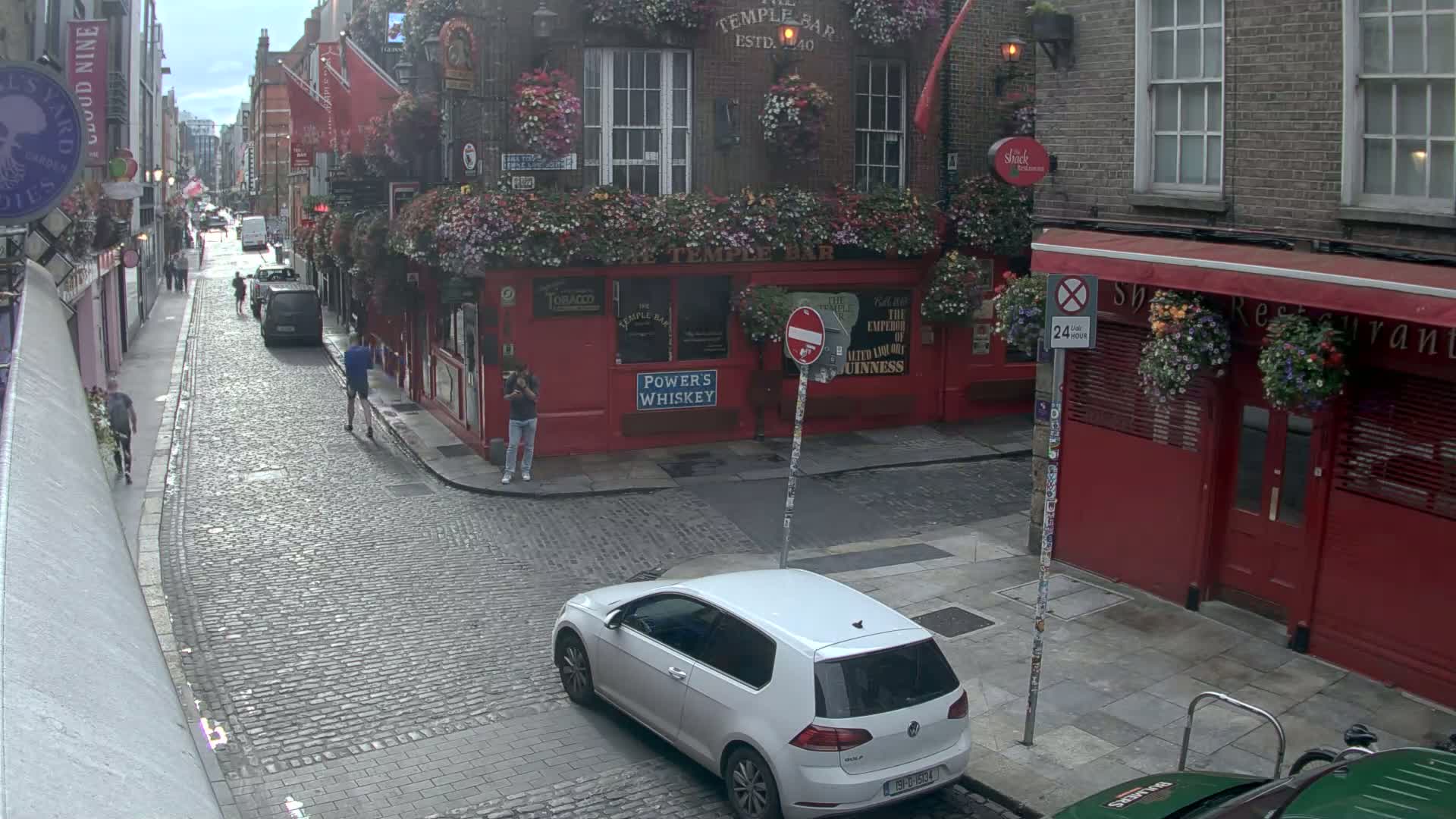 A cobblestone street with pedestrians and a white car parked near a red building with flower boxes, under an overcast sky.
