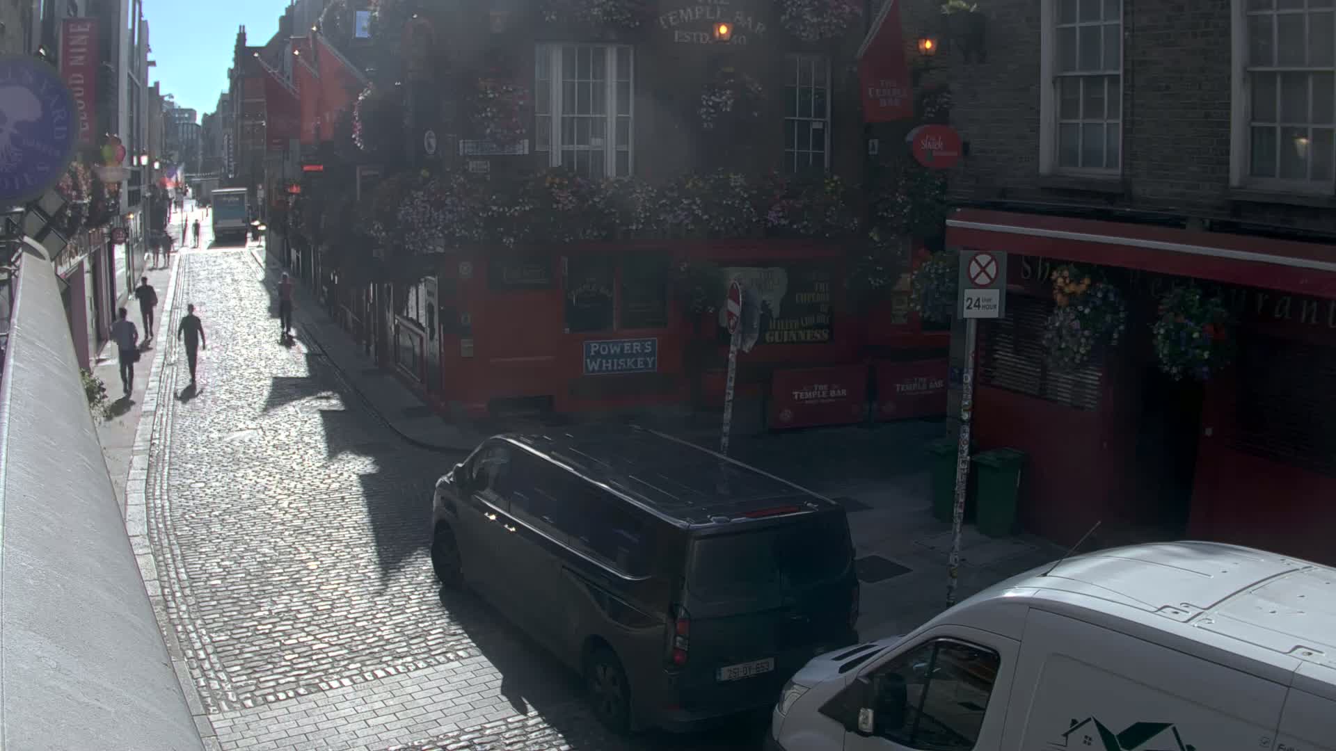 A sunny day on a cobblestone street lined with buildings, with several pedestrians walking and vehicles parked.